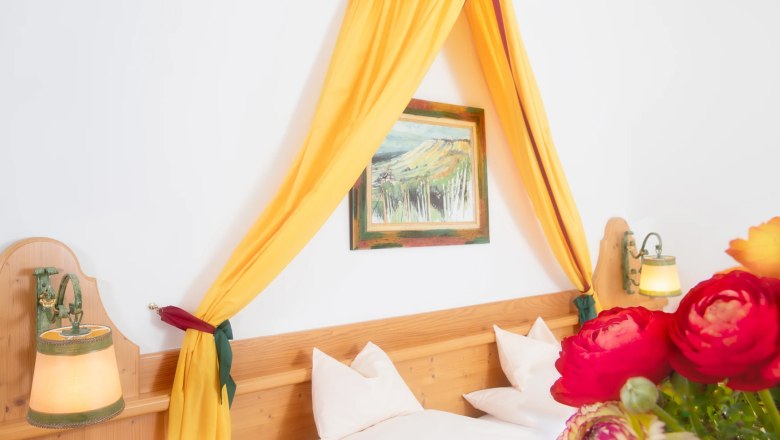 A cozy double room with a yellow canopy, wooden bed, wall lamps and a bouquet of flowers in the foreground.