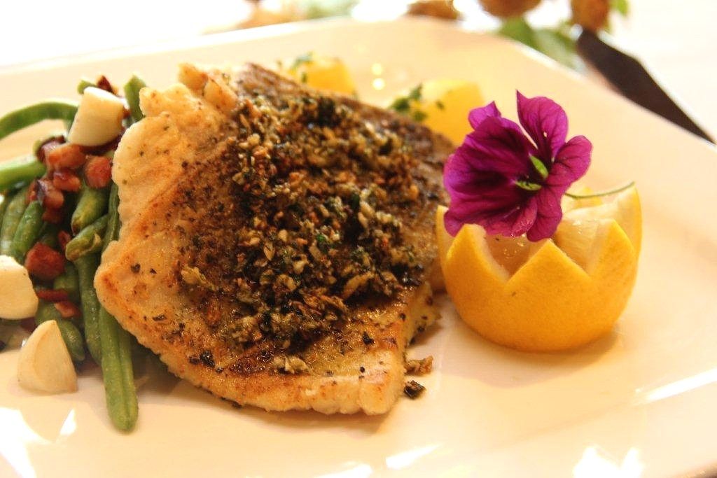 A plate of fried fish fillet, decorated with a flower and a slice of lemon, served with green beans and bacon.