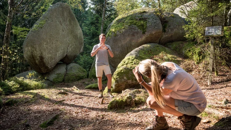 A man poses in front of a large heart-shaped rock in the forest while a woman takes a photo of him.