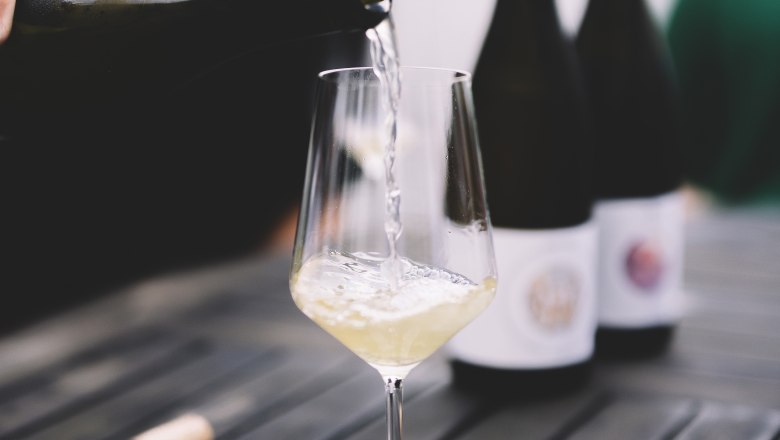 White wine is poured into a glass on a table, with two bottles of wine in the background.