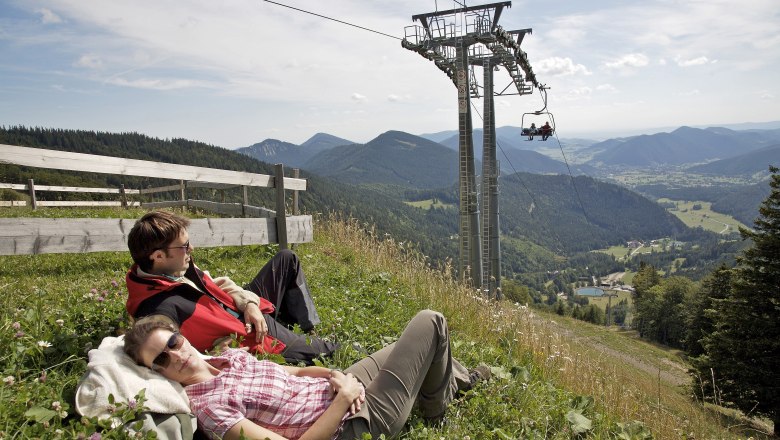 Two people relax on a meadow next to a chairlift with a mountain view.