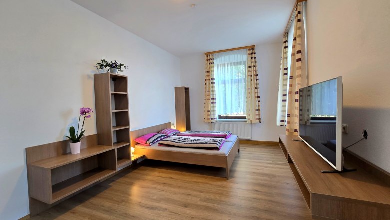 Modern bedroom with wooden floor, double bed, TV and shelves. Large windows with striped curtains.