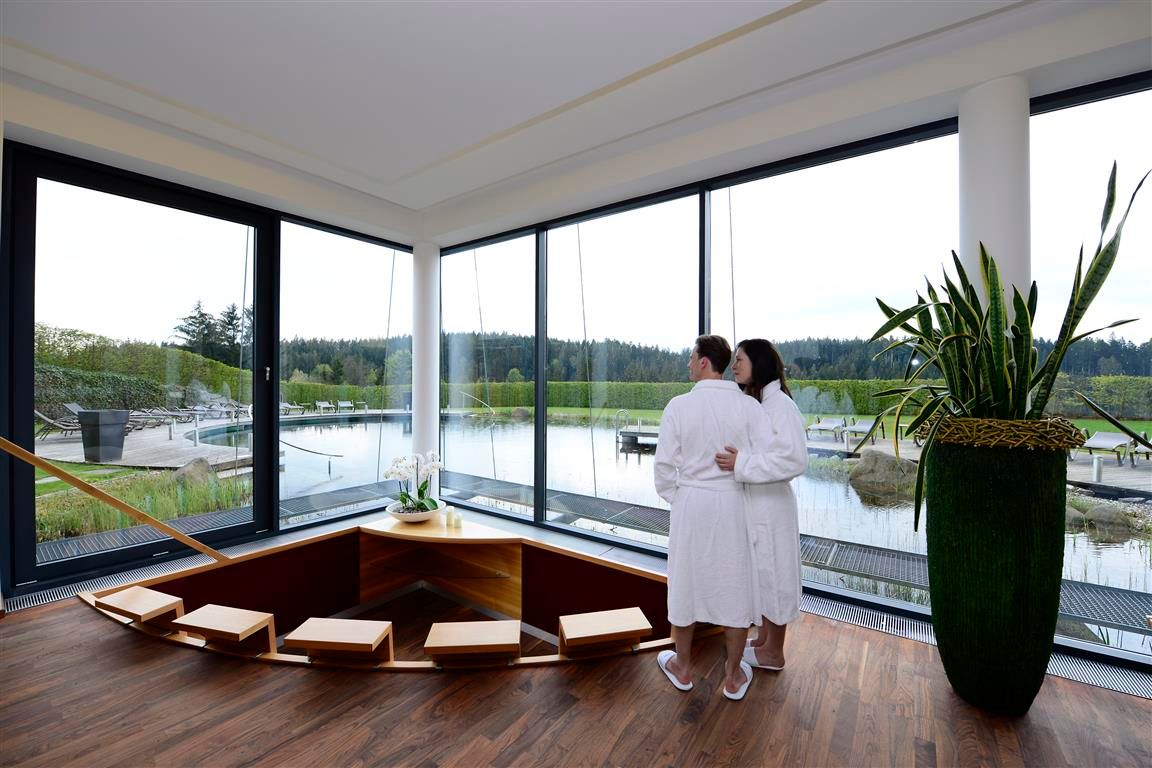 A couple in bathrobes stand in a modern wellness area with a view of a pond.