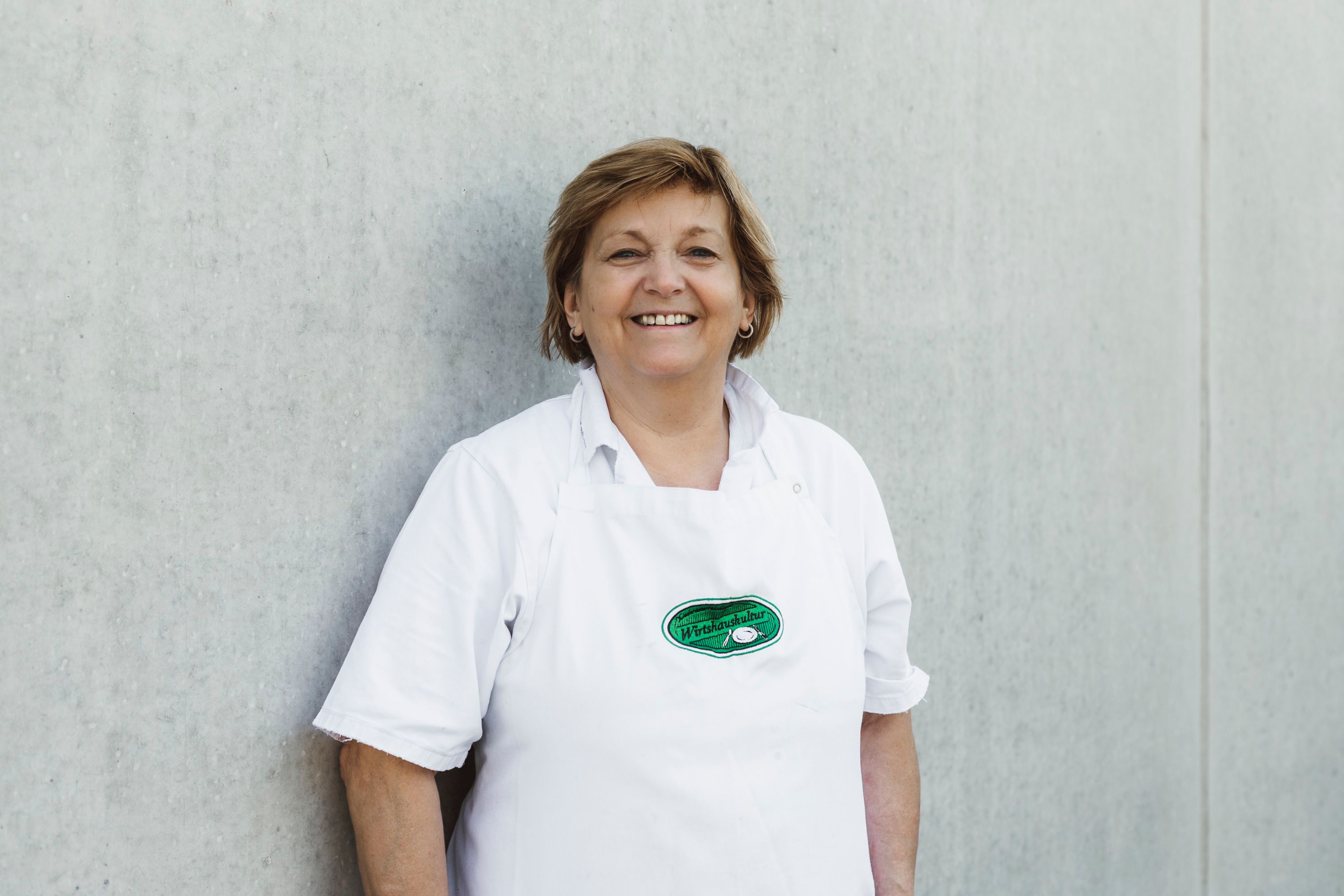 Woman in a white apron in front of a gray wall.