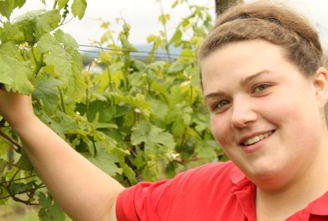Woman in a red shirt stands smiling in a vineyard.