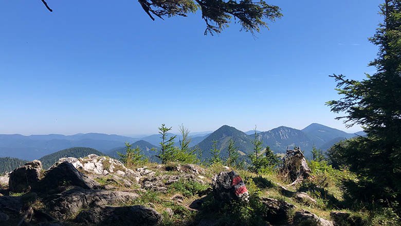 Mountain landscape with rocks and trees in the foreground, blue sky in the background.
