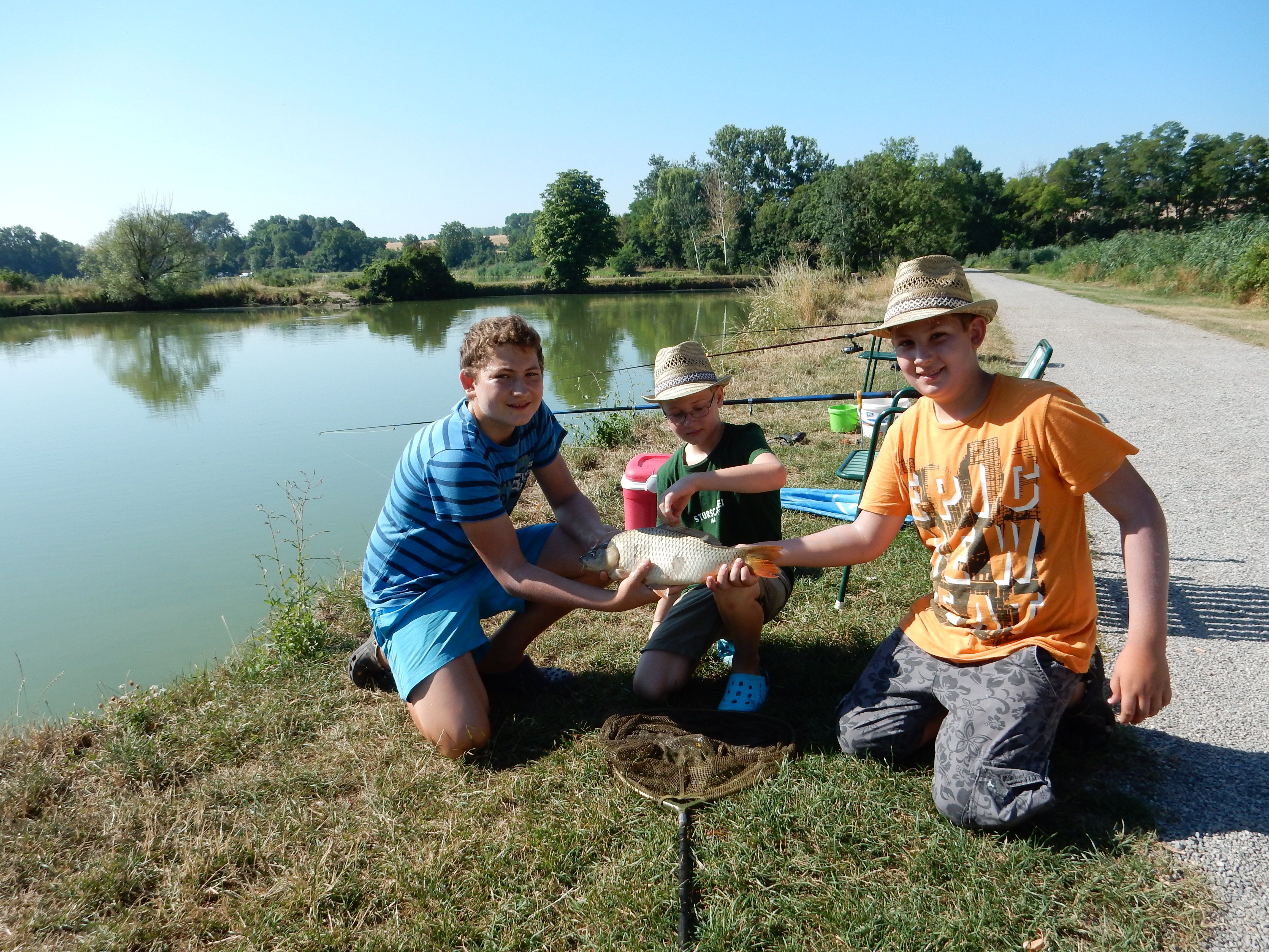 Three boys pose with a caught fish on the shore of a lake.