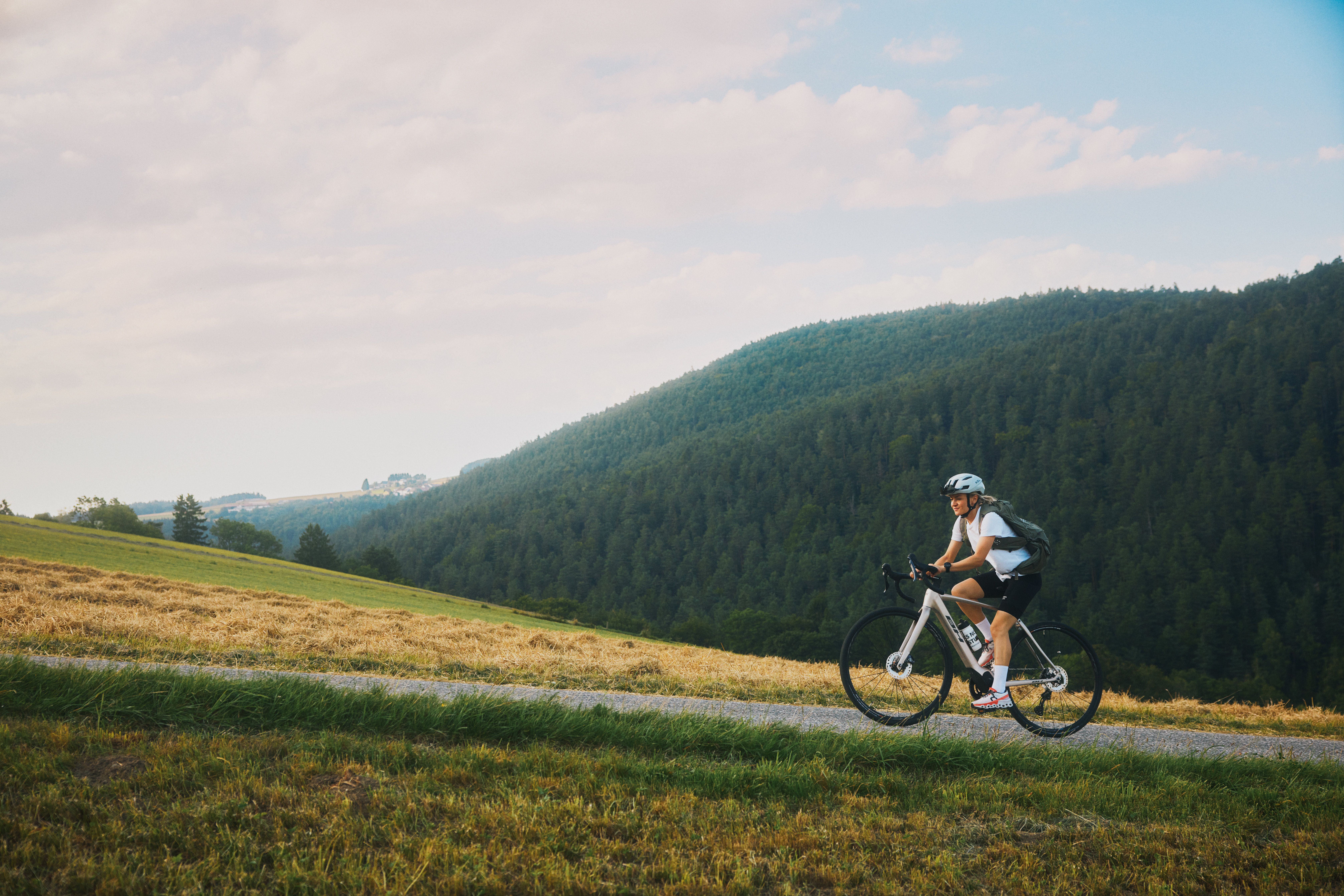A cyclist rides along a narrow, ascending path, with wooded hills in the background.