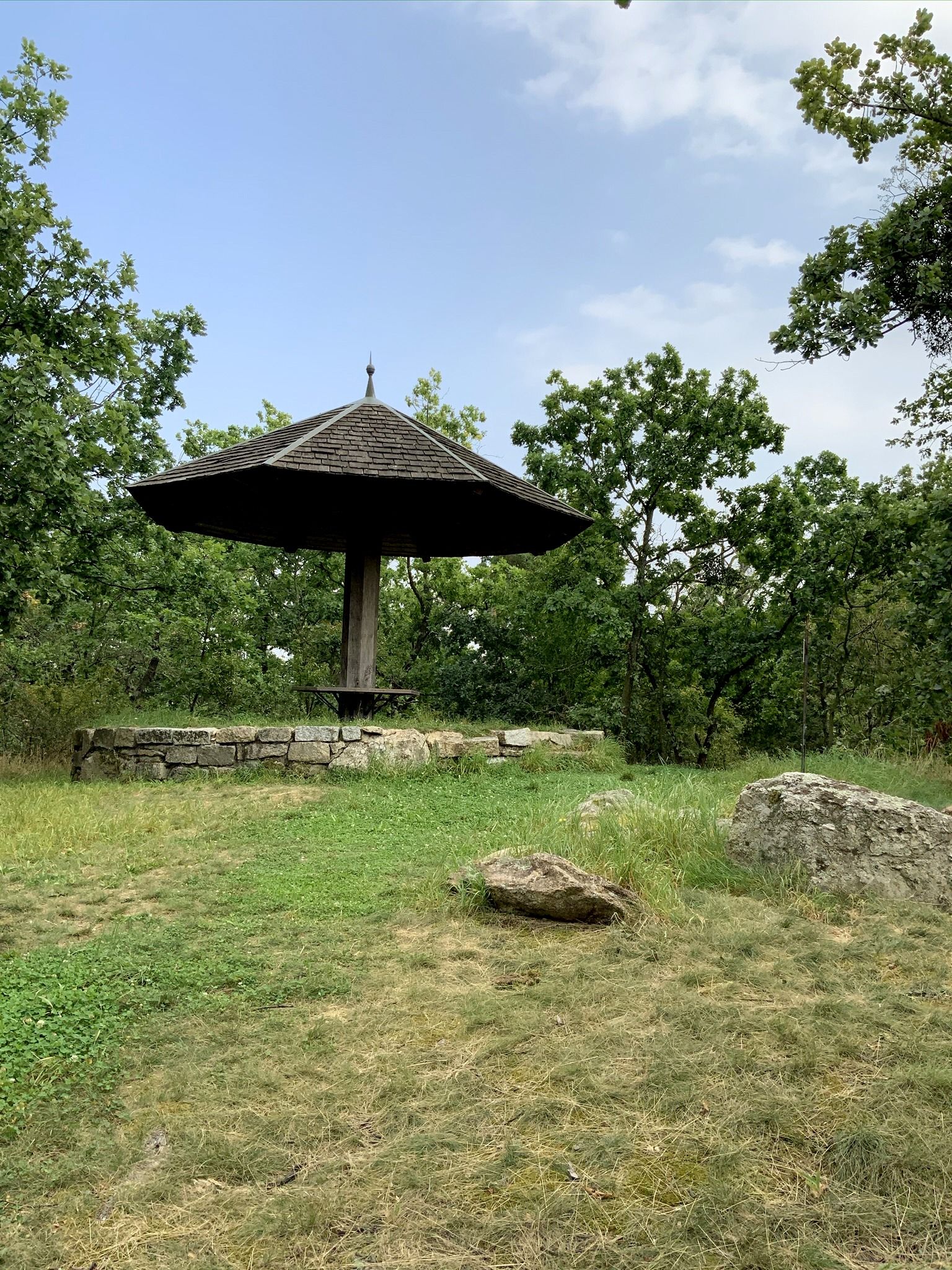 A wooden pavilion with a pointed roof in a green, wooded setting.