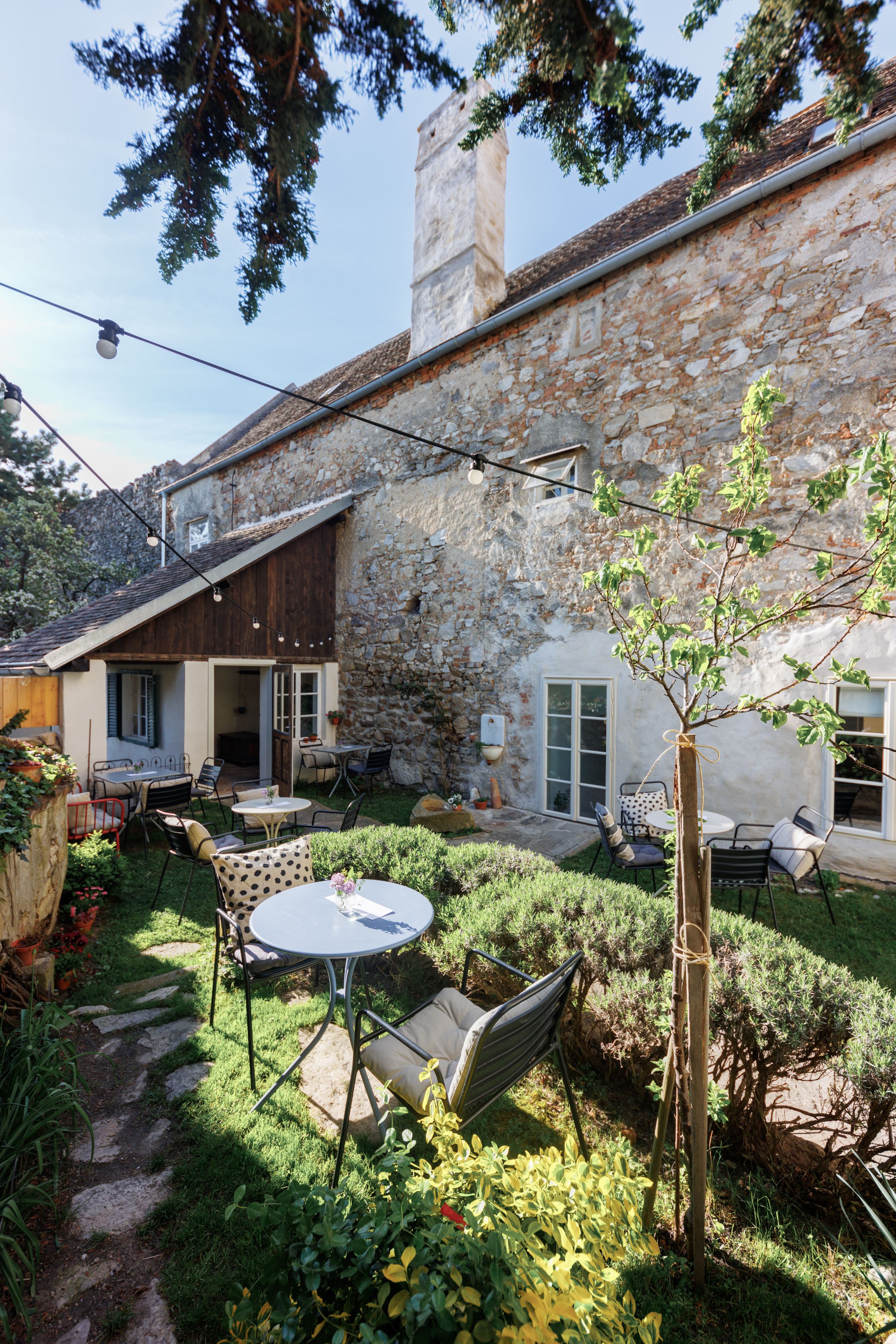 A cozy garden with tables and chairs, surrounded by plants and an old stone wall. Fairy lights hang over the garden.