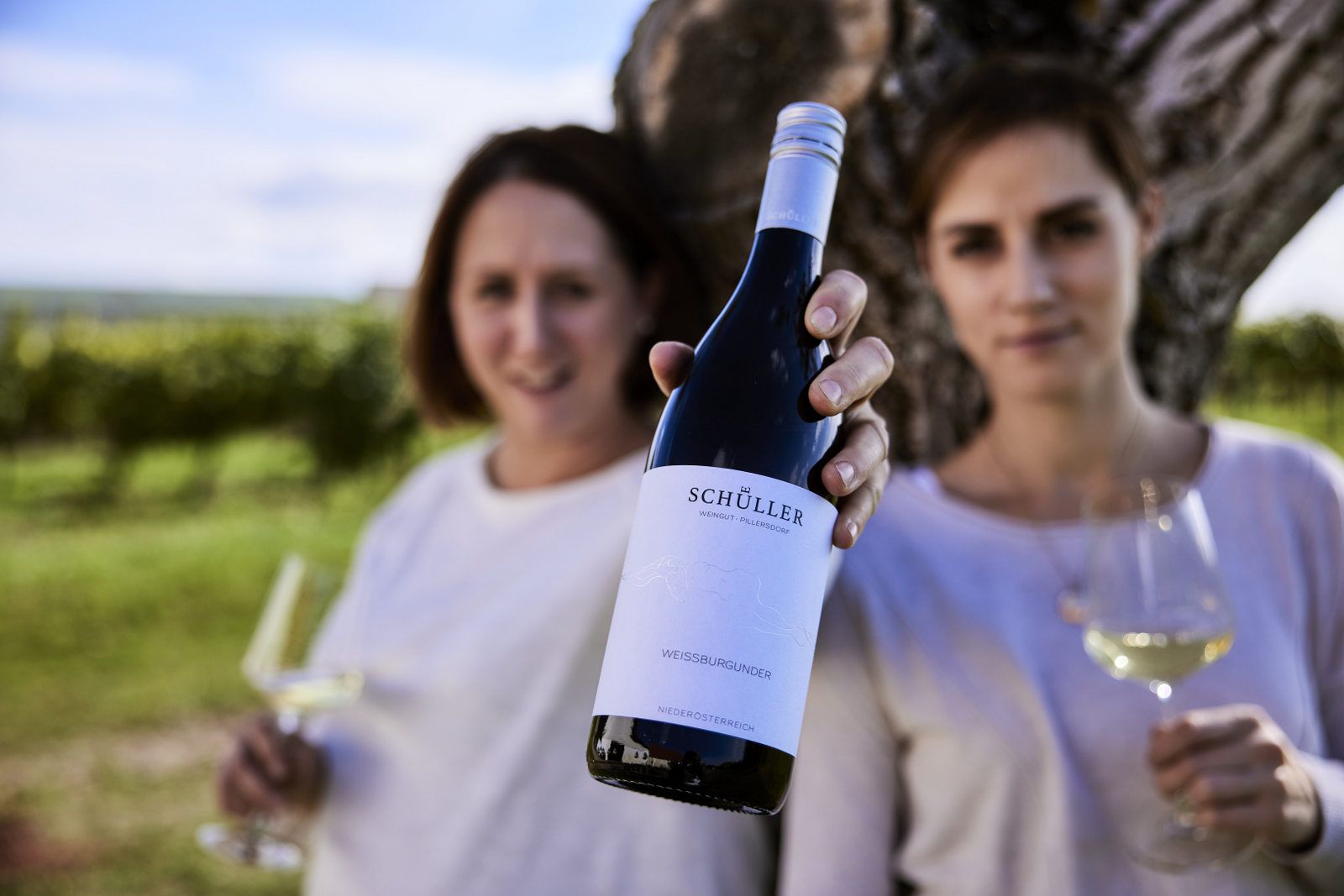 Two people holding a bottle of Schüller Pinot Blanc and wine glasses in front of a tree in a vineyard.
