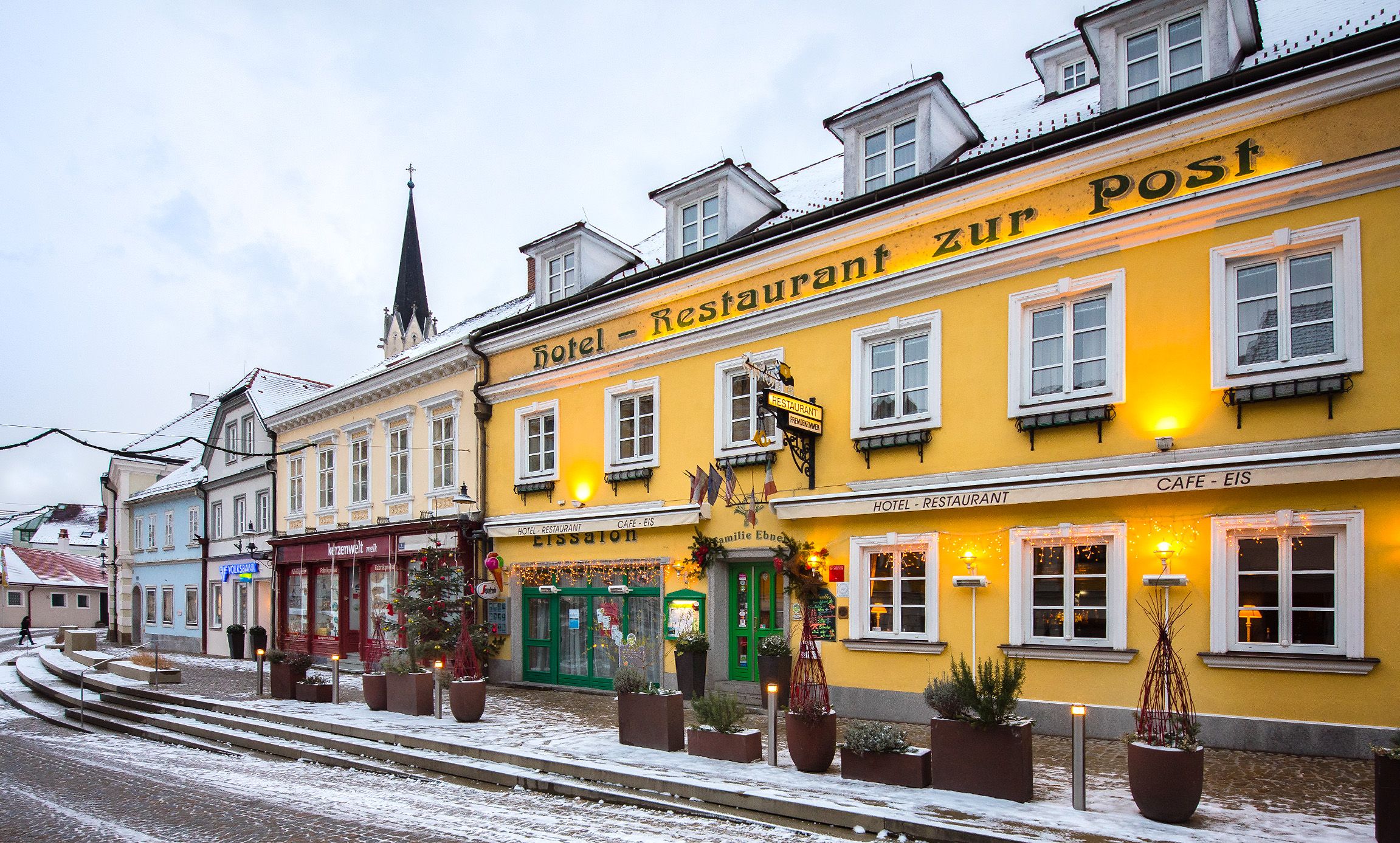 Exterior view of a yellow building with the inscription 'Hotel-Restaurant zur Post' in winter, surrounded by snow and decorated with lights.