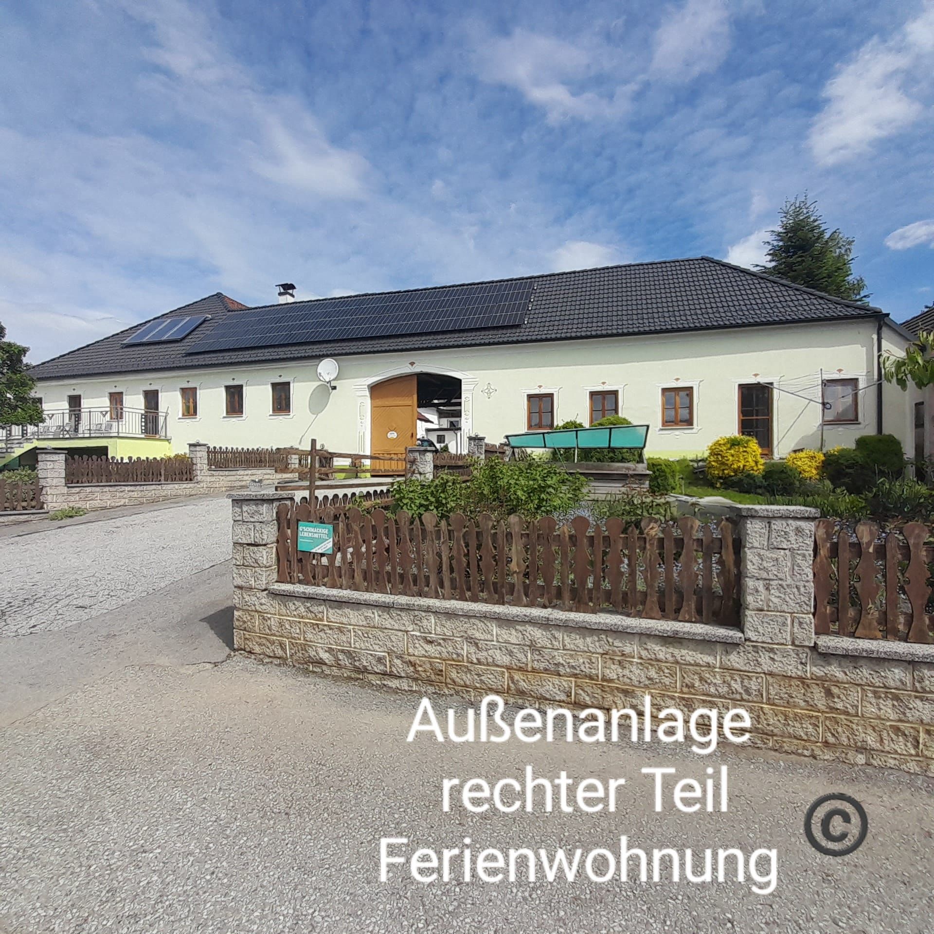 A large building with a yellow façade and black roof, surrounded by a garden and a wooden fence. The sky is blue with a few clouds.