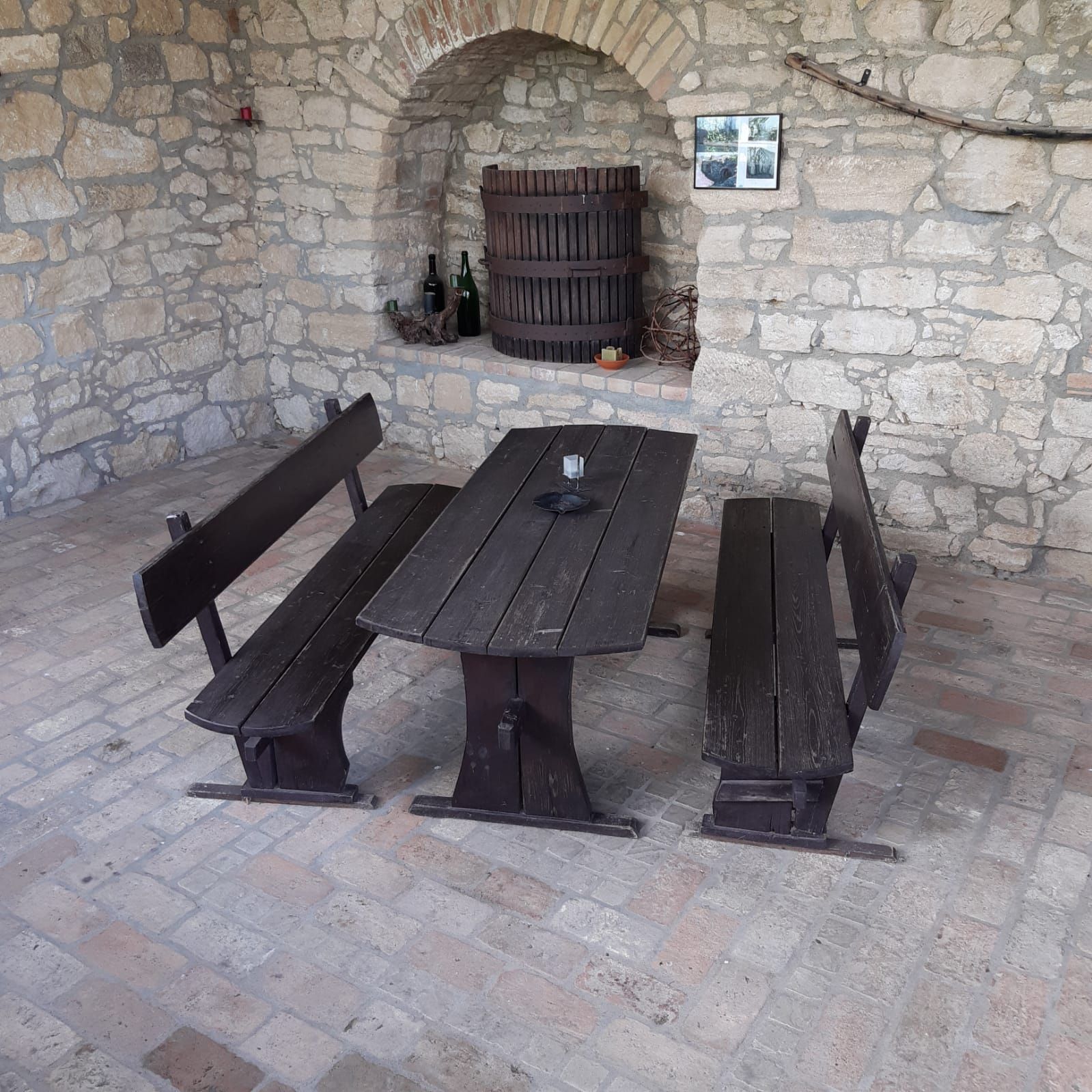 A rustic seating area with a wooden table and benches in front of a stone wall with a wine barrel.