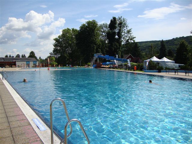 A large outdoor pool with clear water, surrounded by trees and hills under a blue sky.