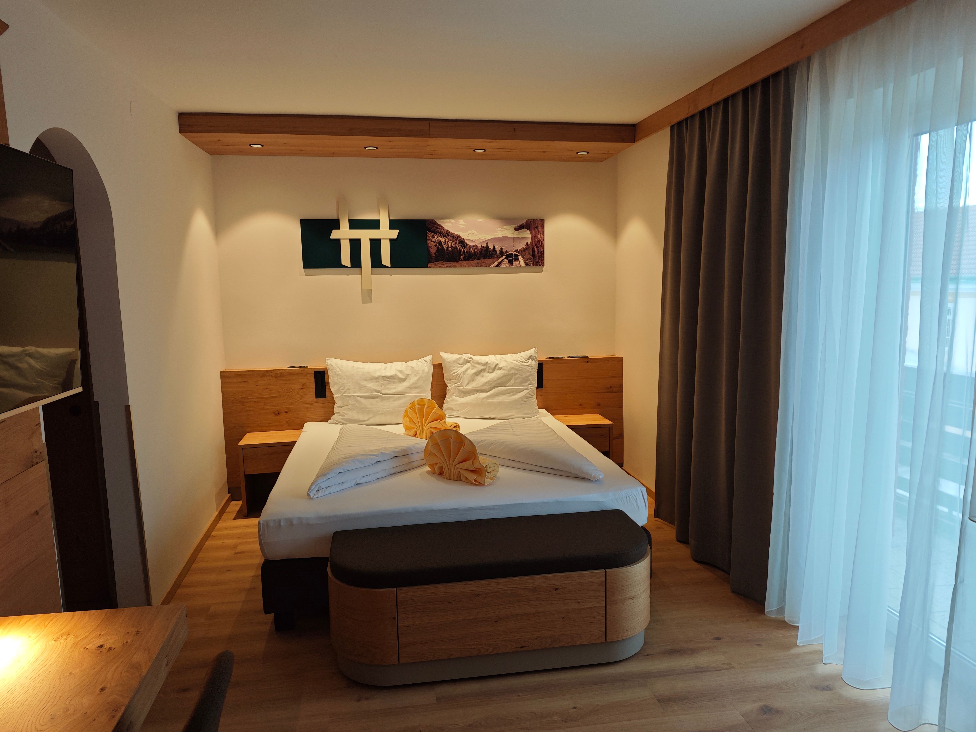 A modern double room with a large bed, wooden furniture, and a large window with curtains.