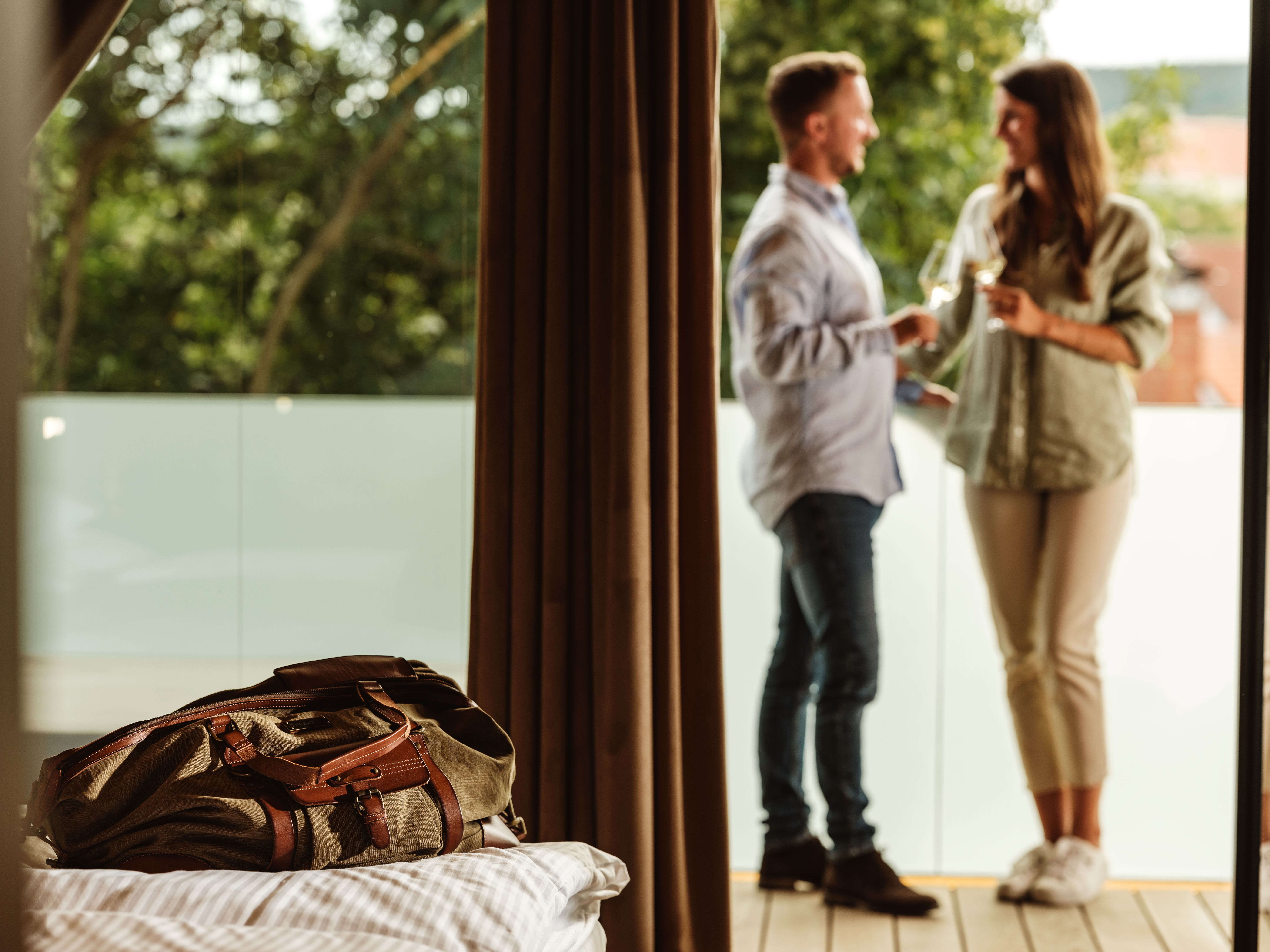 A couple is standing on a balcony, clinking glasses; a travel bag lies on a bed in the foreground.