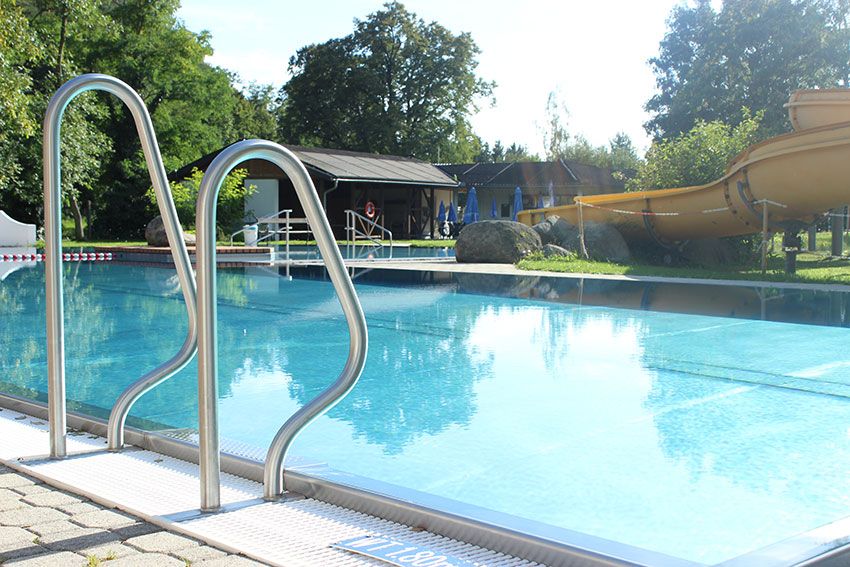 An outdoor pool with slide and ladder, surrounded by trees and a building in the background.