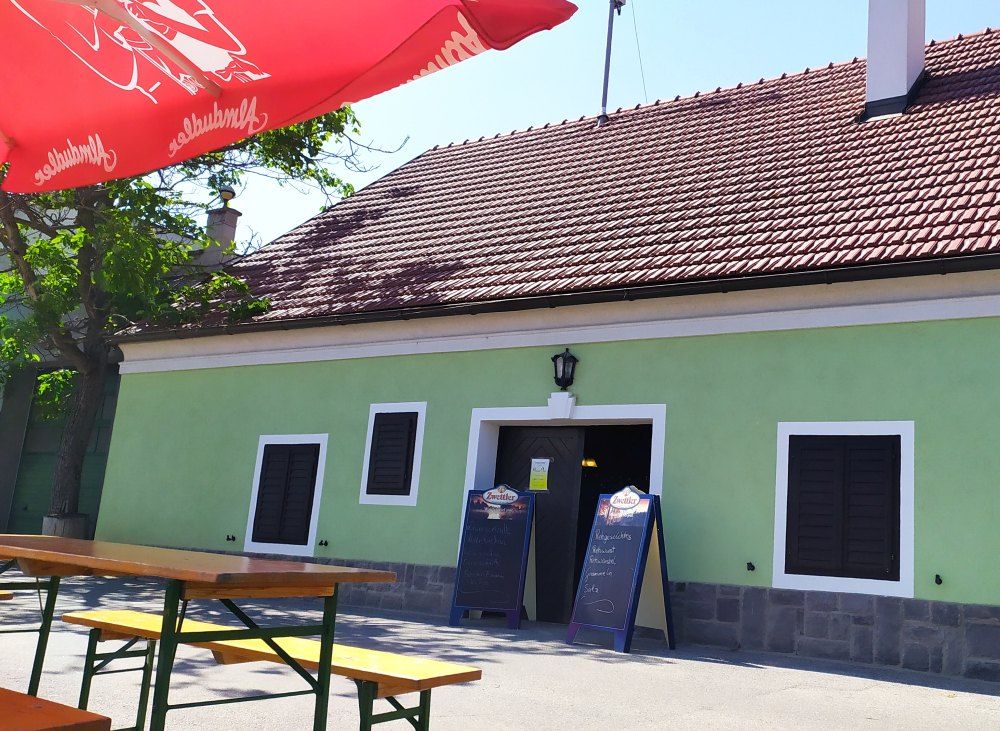 Exterior view of a traditional wine tavern with green paint and red roof tiles. There are wooden benches and a red parasol in front of the building.