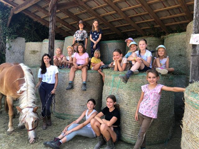 A group of children are sitting on hay bales in a stable, with a horse standing next to them. The children are smiling and wearing casual clothes.