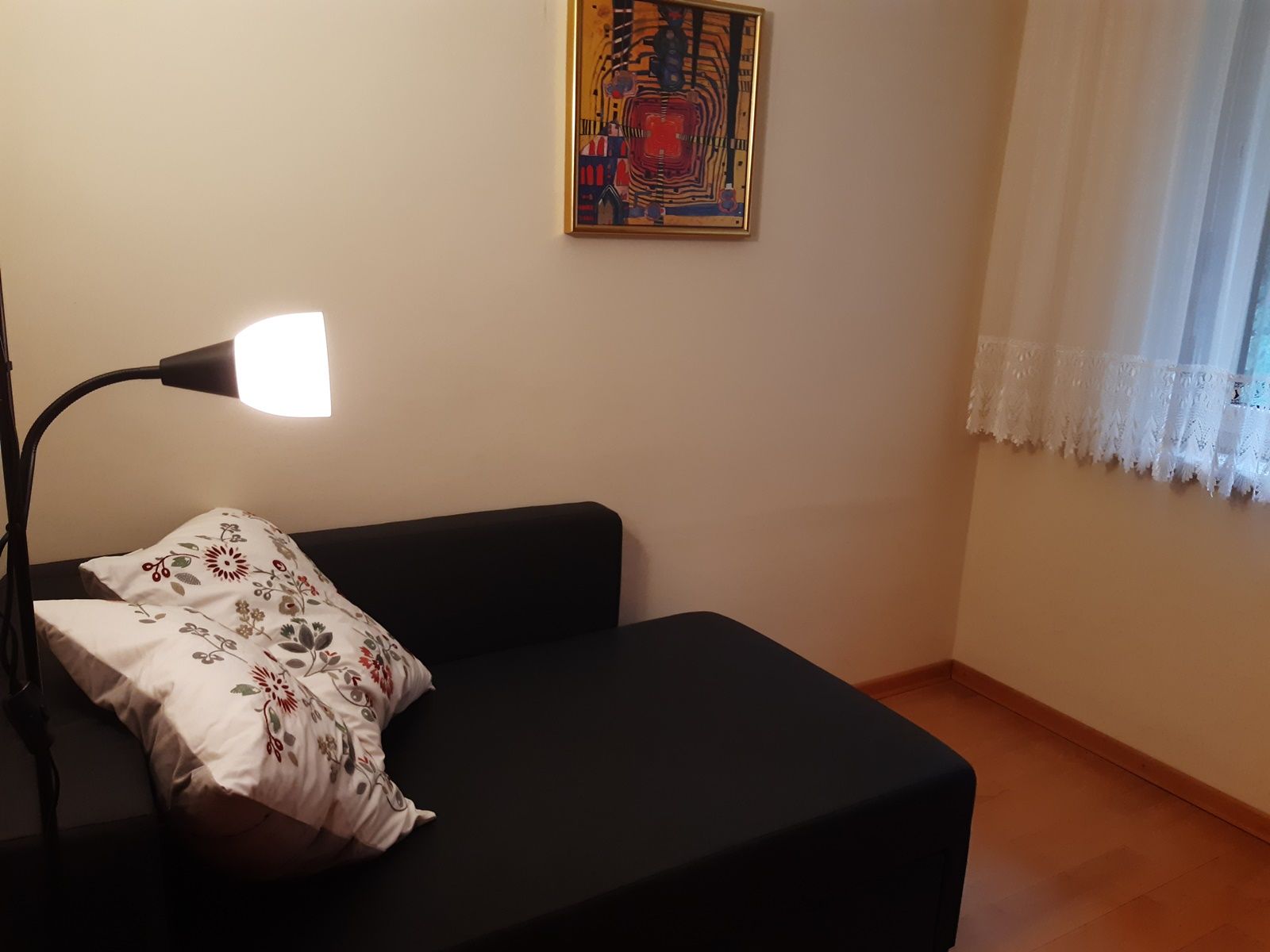Cozy reading corner with black sofa, floor lamp and decorative cushion. A colorful picture hangs on the wall, next to it a window with curtains.
