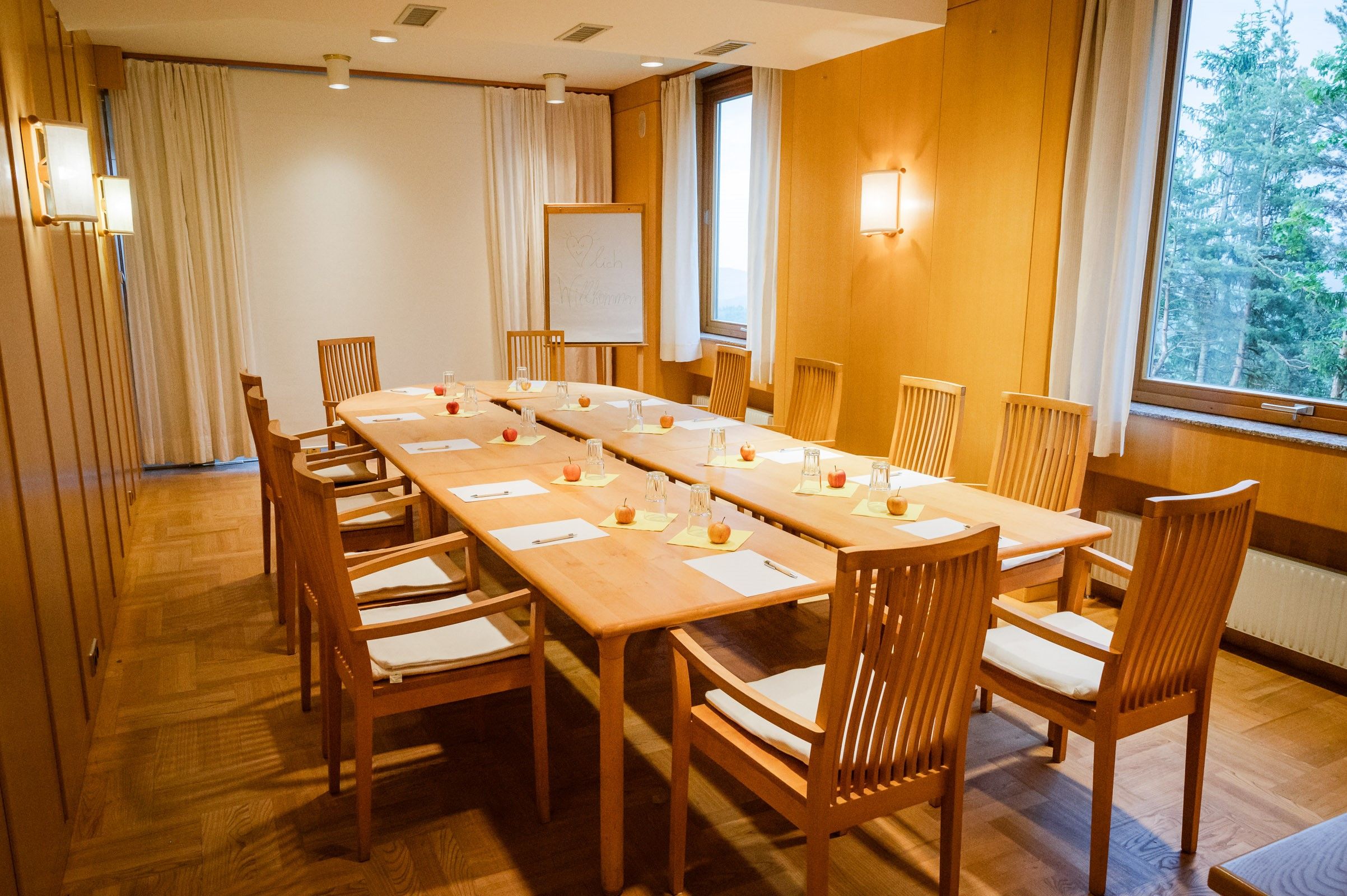 A seminar room with a long wooden table surrounded by chairs. There are notepads, pens and apples on the table.