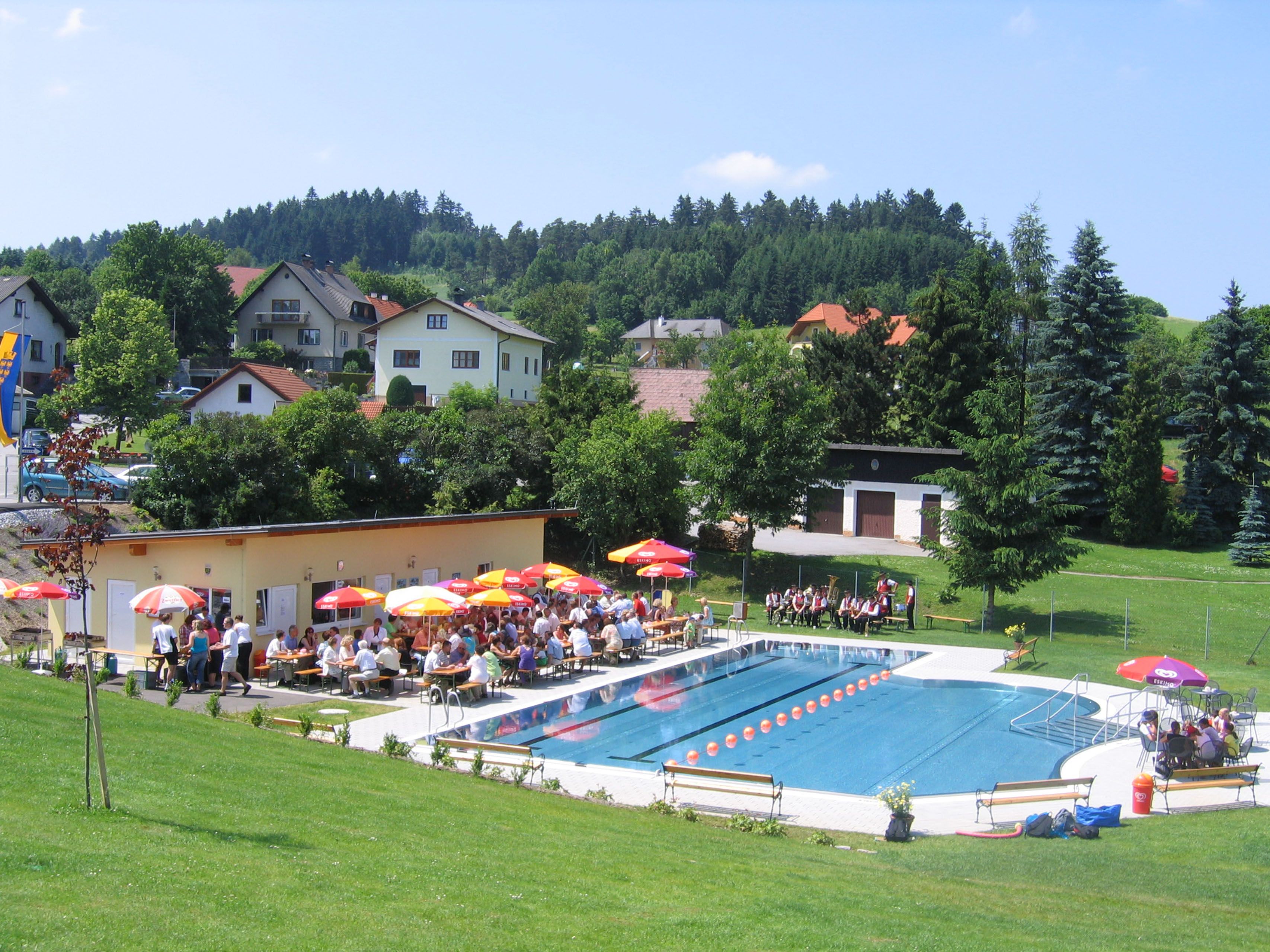Outdoor pool with swimming pool, sunshades and people on deckchairs, surrounded by green hills and houses.