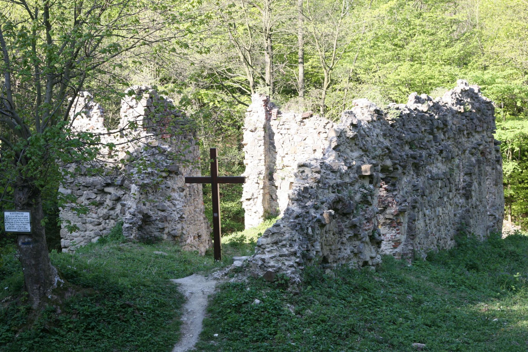Ruins of an old monastery with a wooden cross in the foreground, surrounded by trees.