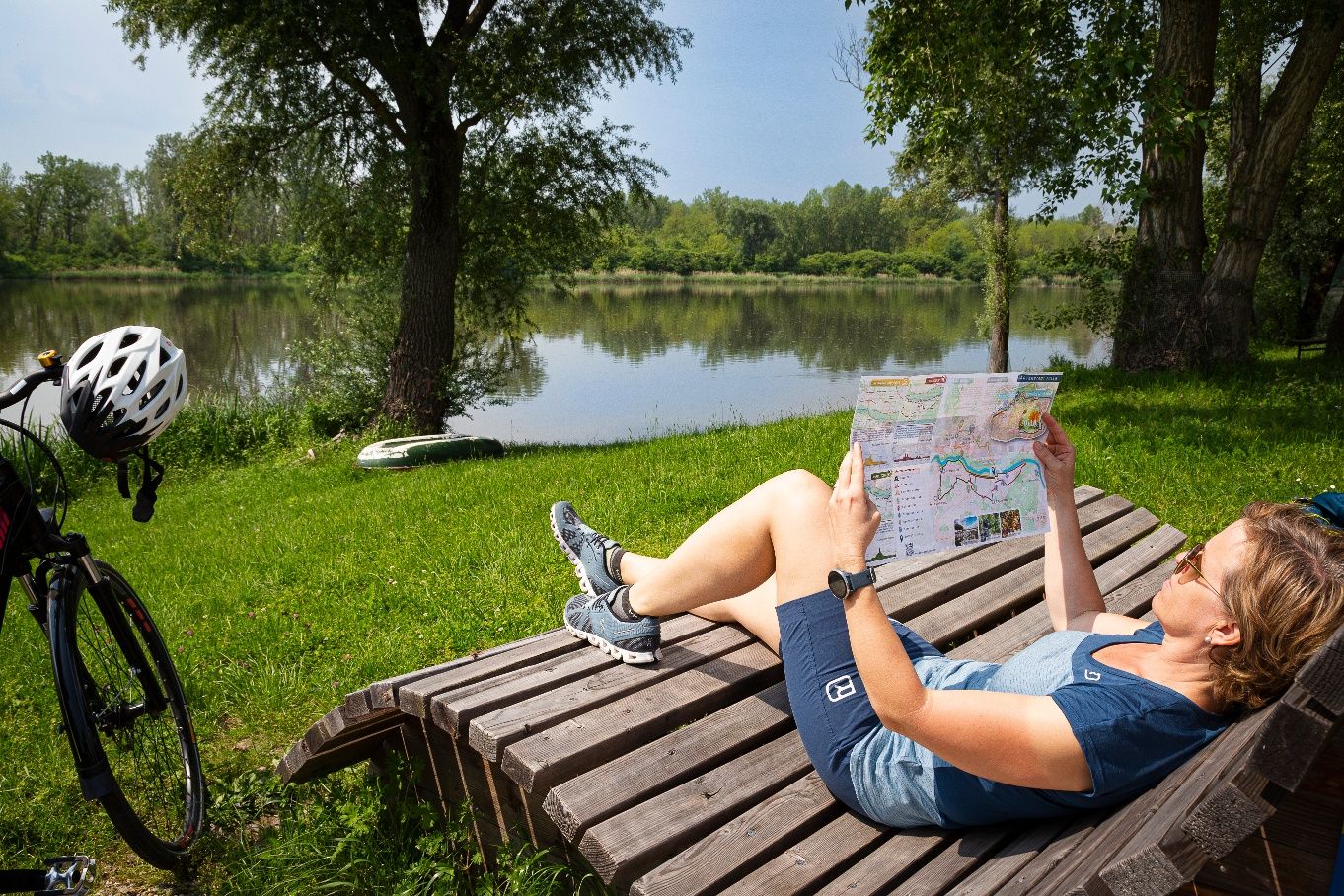 A woman is lying on a wooden bench by the lake reading a map. A bicycle with a helmet stands next to her.