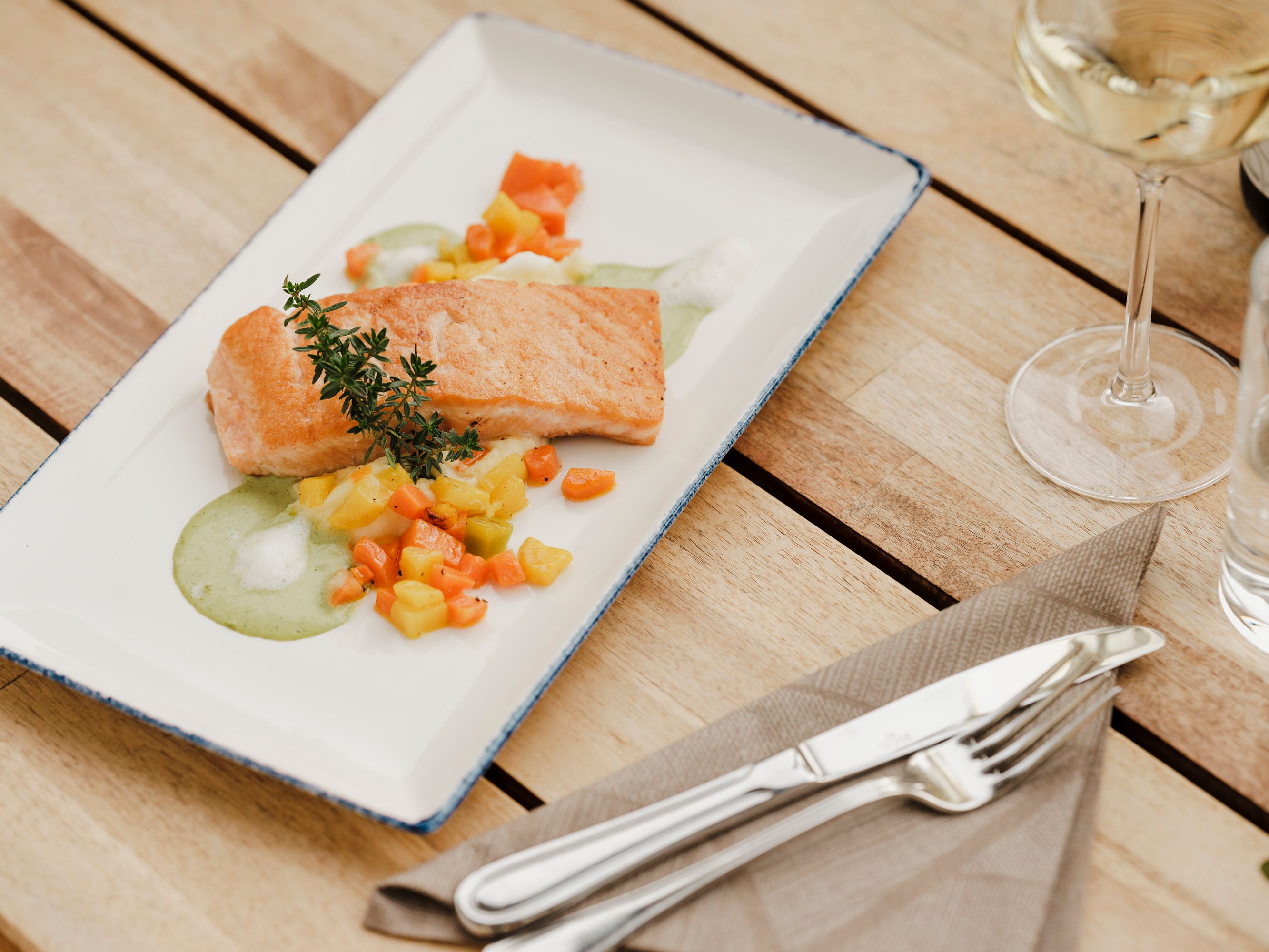 A plate with roasted salmon on vegetables and green sauce, decorated with a sprig of herbs, on a wooden table with cutlery and a wine glass.