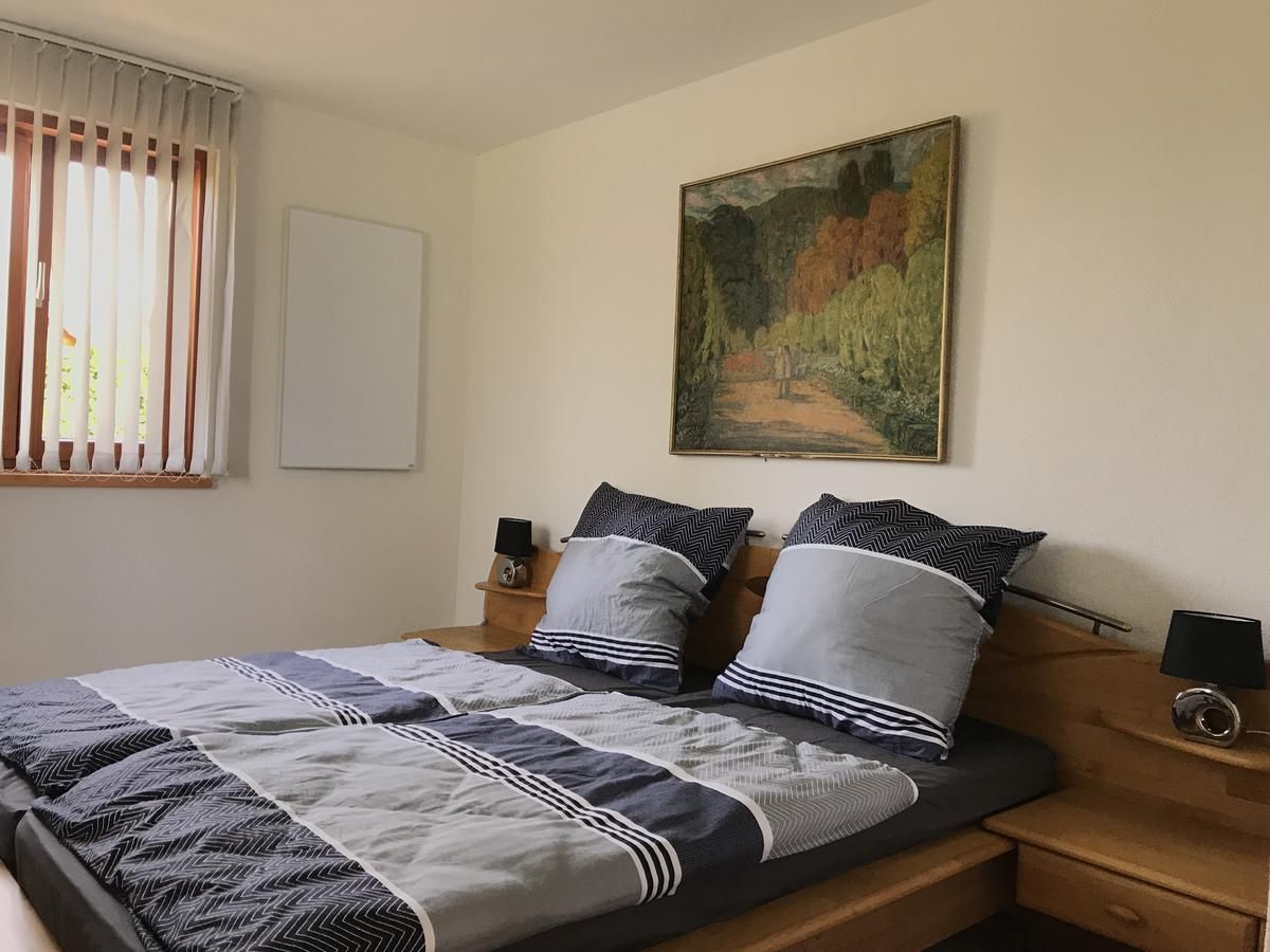 A cozy bedroom with a double bed, grey bed linen, a painting on the wall and a window with blinds.