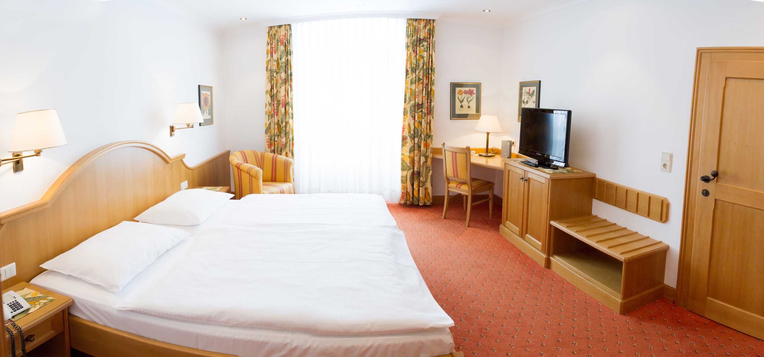 A hotel room with a double bed, desk, TV and armchair. Bright curtains and floral drapes adorn the window.