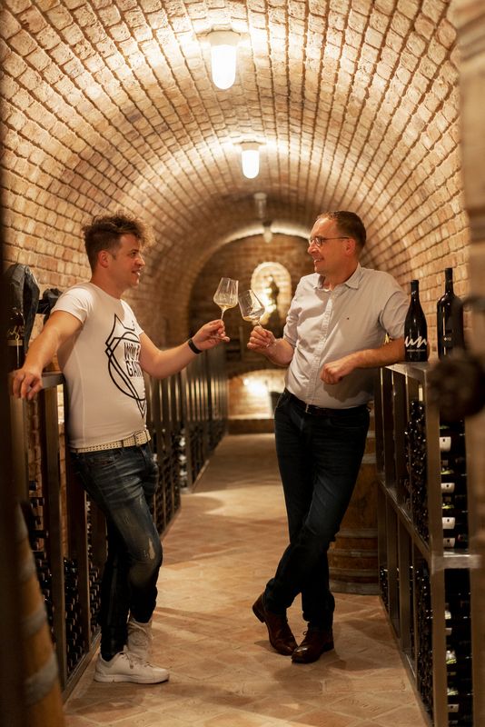 Two men are standing in a wine cellar, clinking glasses of wine.