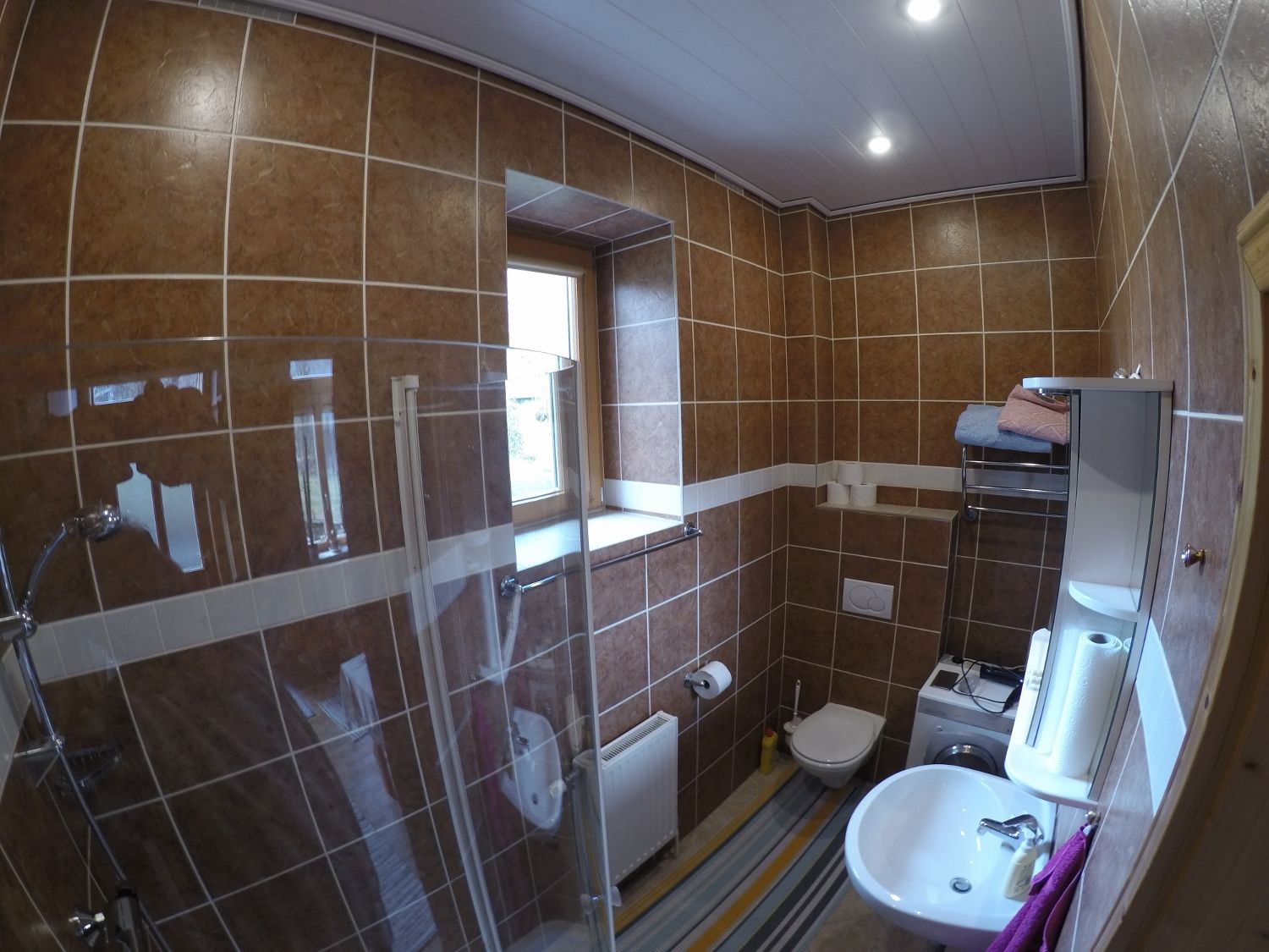 Bathroom with brown tiles, shower, washbasin, toilet and washing machine.
