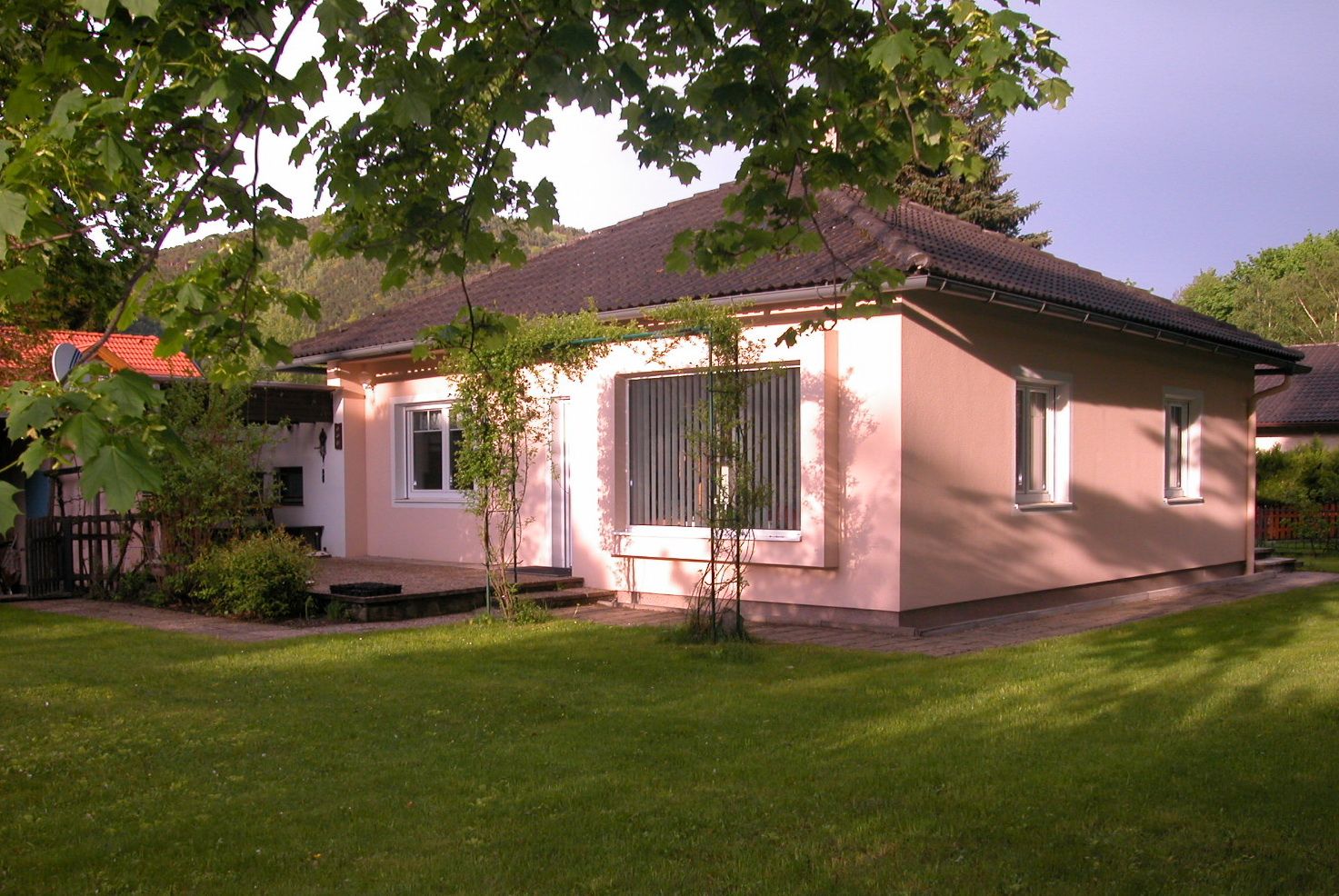 A pink-painted vacation home with a dark roof, surrounded by a green garden and trees.