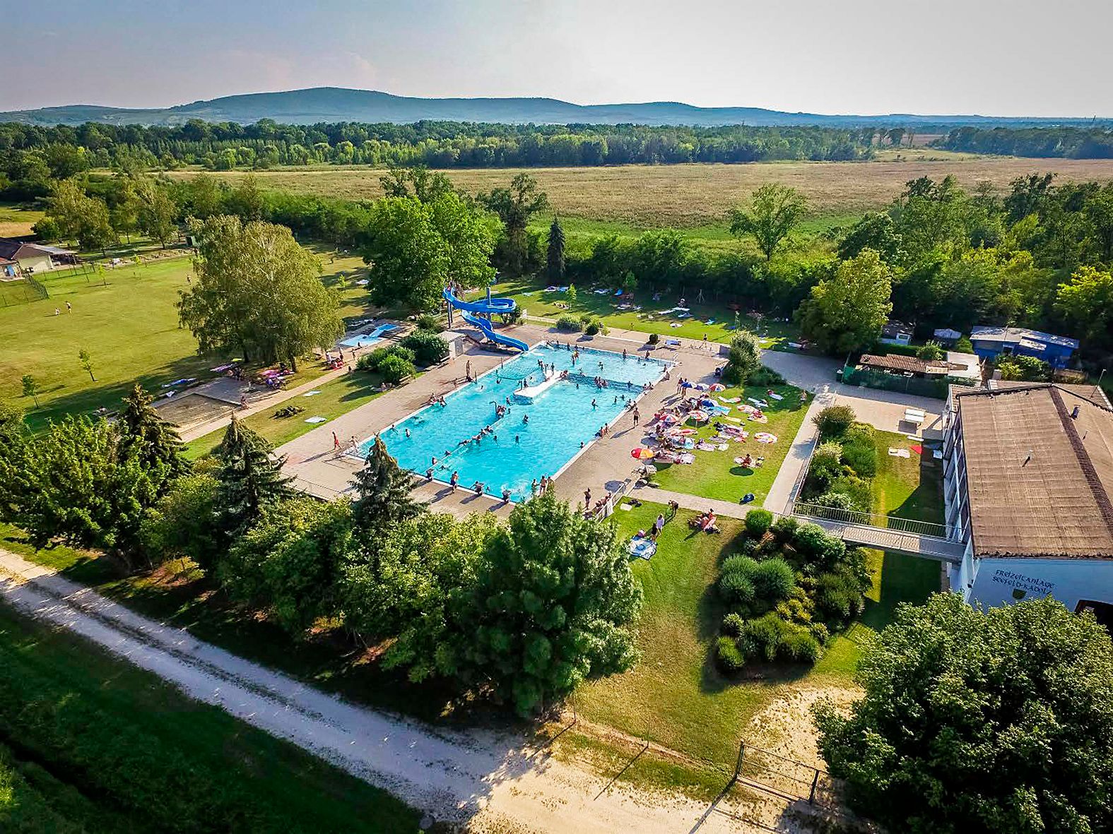 Aerial view of an outdoor pool with swimming pool, slide and sunbathing area, surrounded by trees and fields.