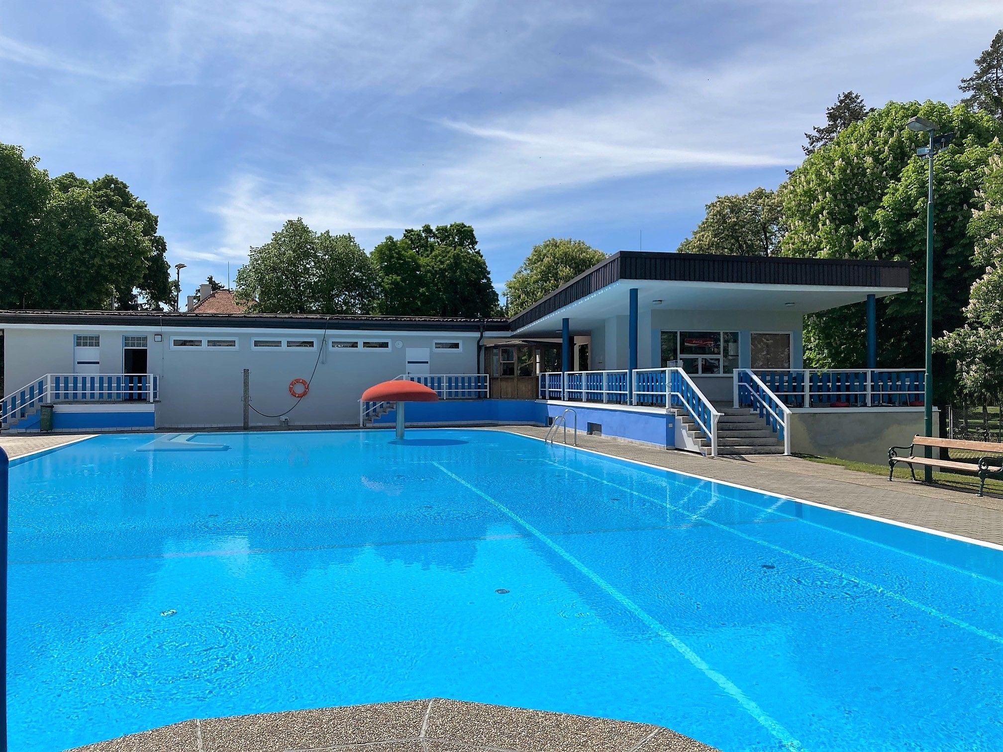 An outdoor pool with clear water, surrounded by trees and a building with blue railings.
