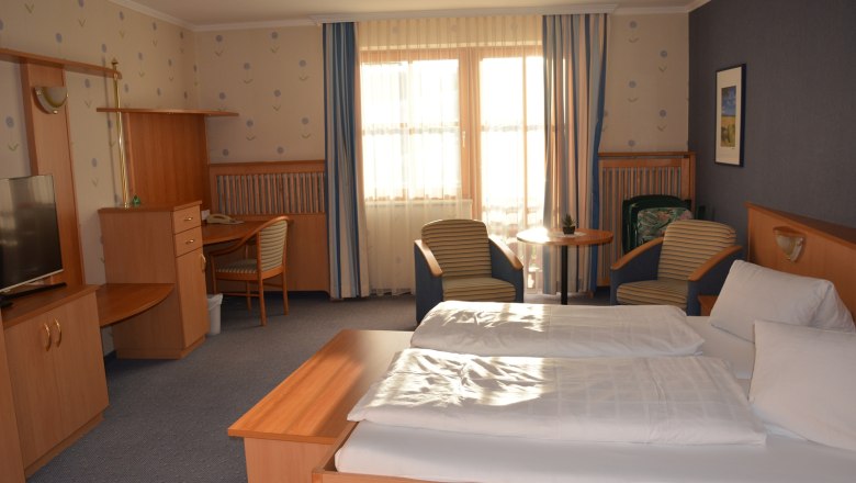 A cozy hotel room with a double bed, desk, TV and two armchairs in front of a window with curtains.