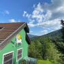 Green house with unicorn picture on the wall, surrounded by trees and mountains under a blue sky.