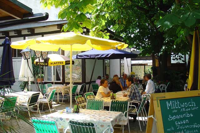 A cozy garden with tables, chairs and parasols, surrounded by trees. People are sitting and chatting.