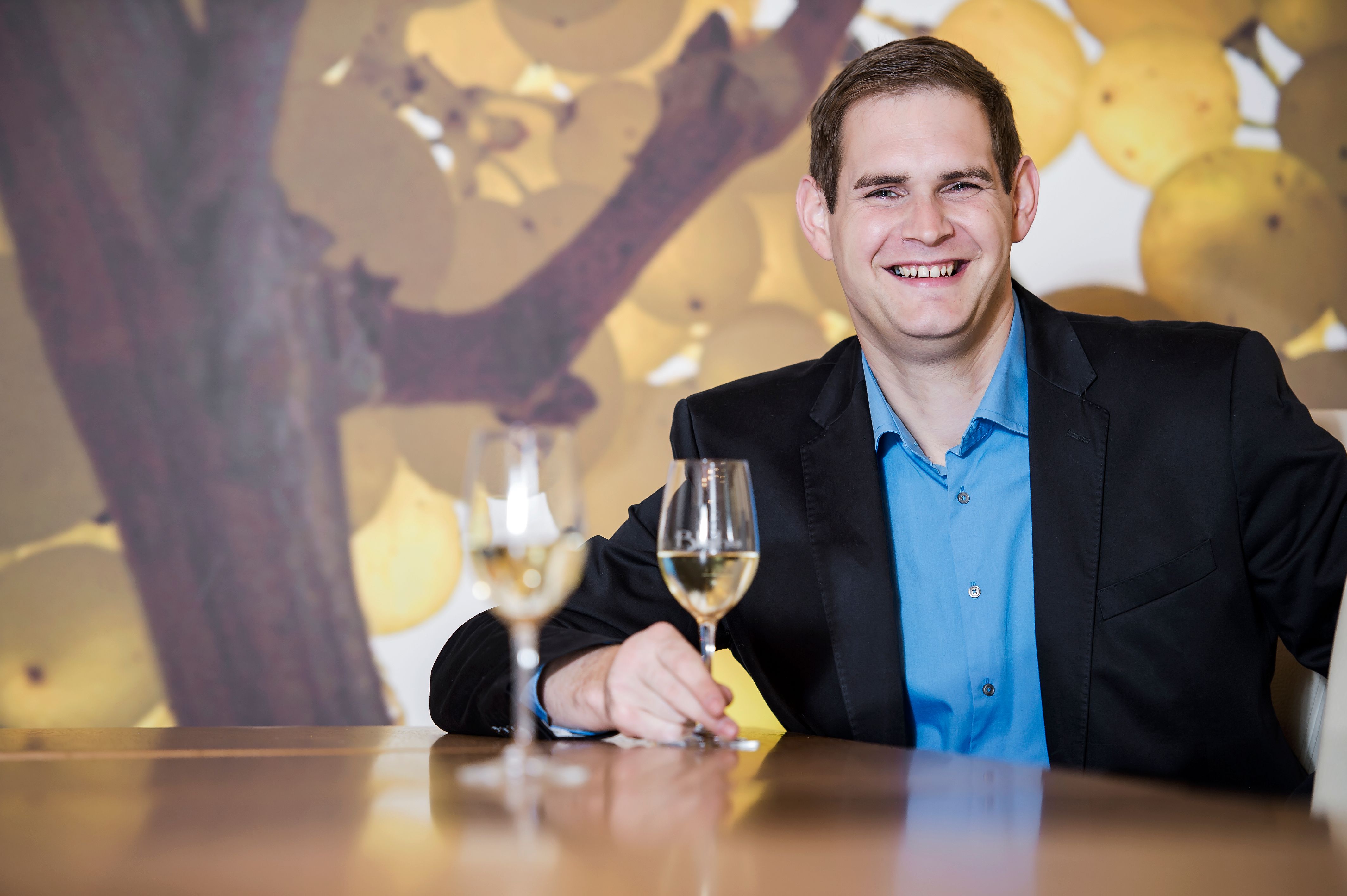 A man in a suit sits at a table with two wine glasses in front of a background of grapes.