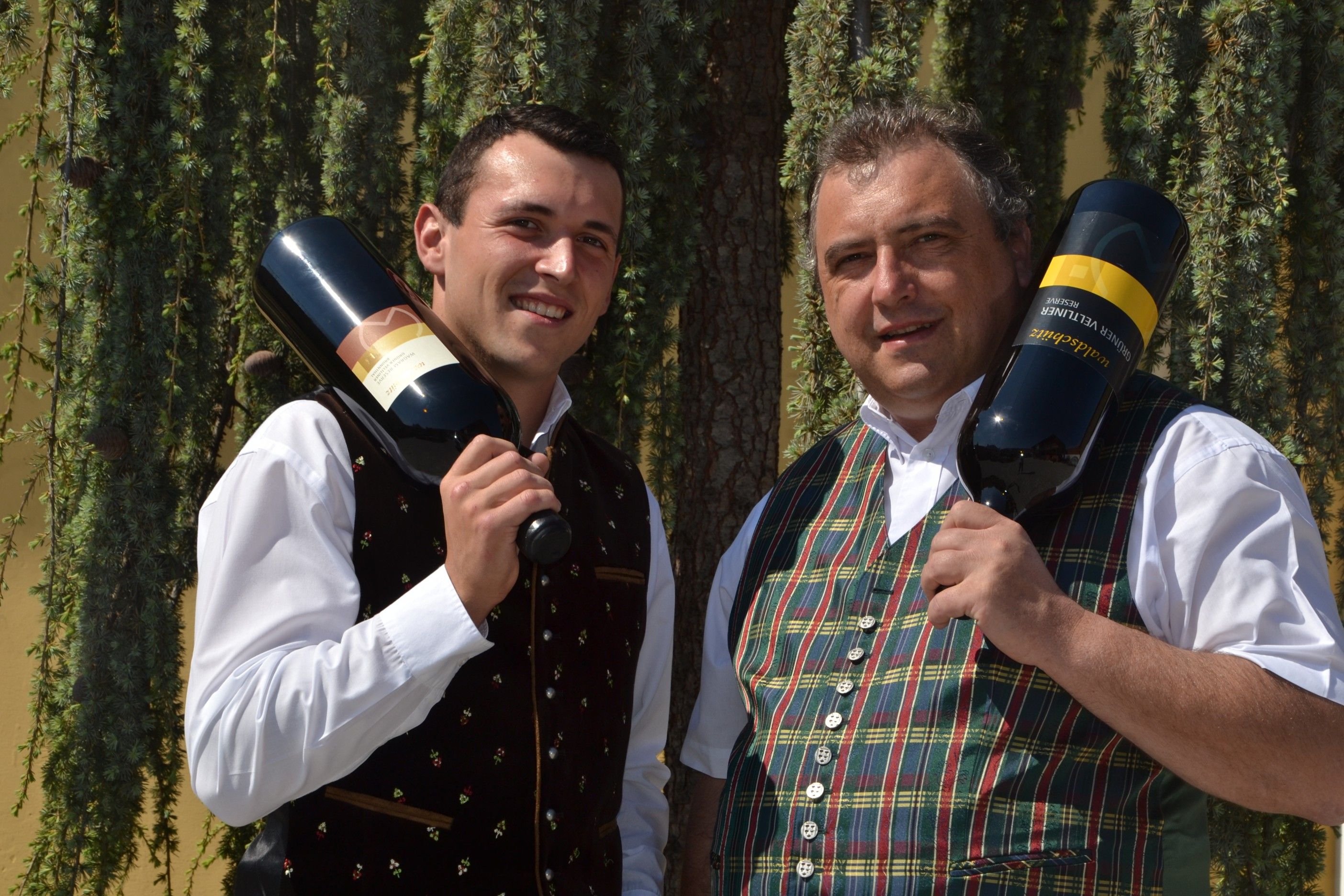 Two men in traditional dress hold large bottles of wine in front of a tree.