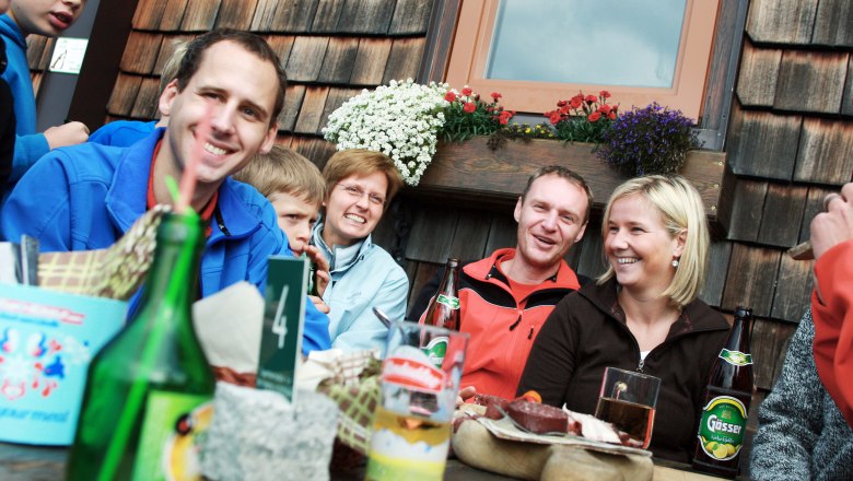 Group of people sitting laughing at a table in front of a wooden hut with flowers.