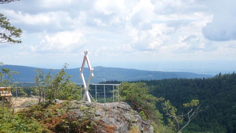 Viewpoint on a mountain with a white, cross-shaped frame and a view of wooded hills under a cloudy sky.
