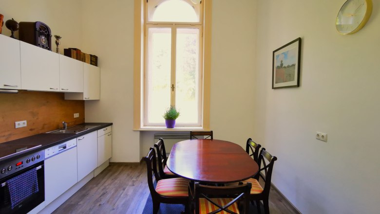 Modern kitchen with dining table and chairs, large window, white cupboards and wooden floor.
