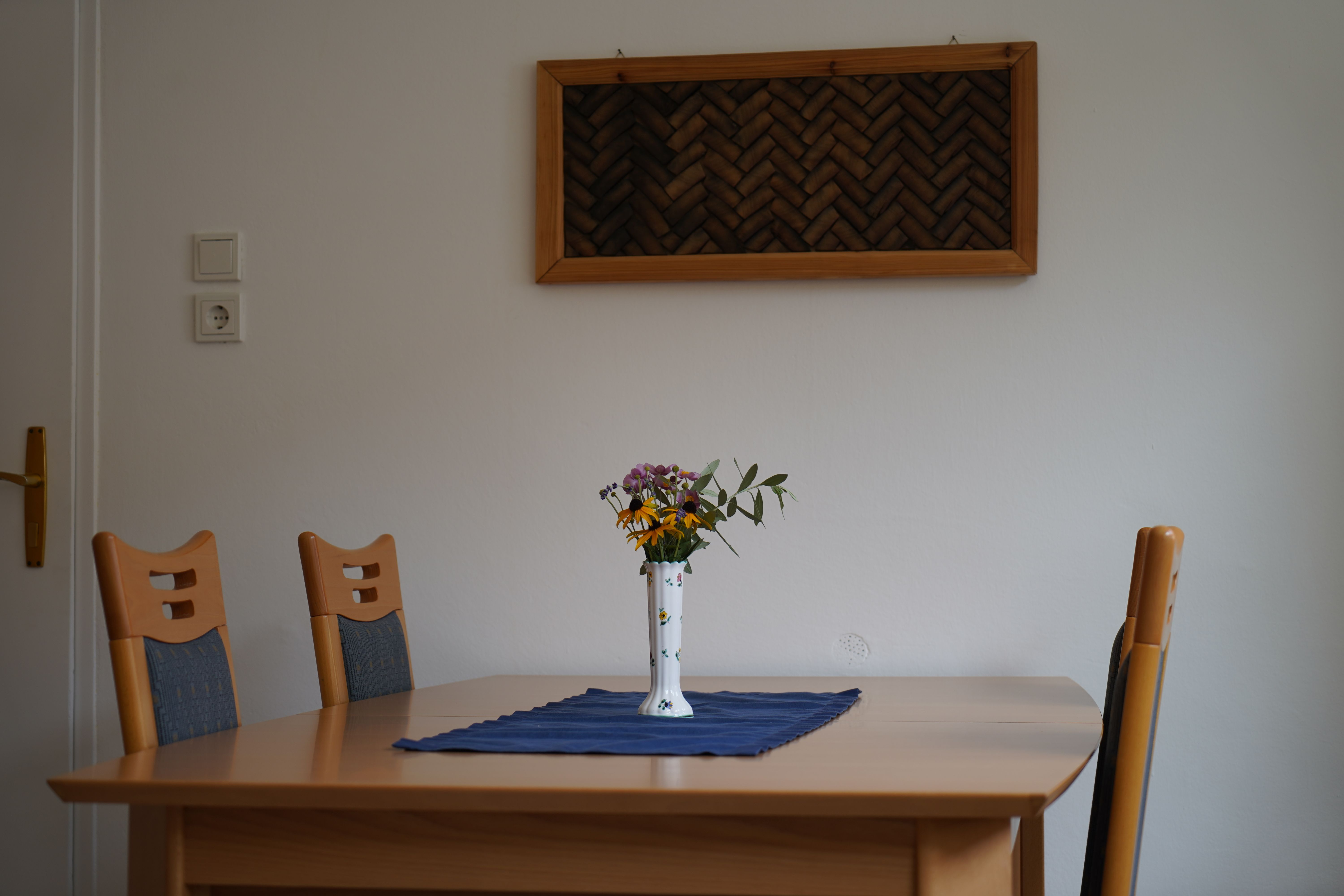 A table with four chairs, a vase of flowers and a blue cloth in a room with a white wall and wall decor.