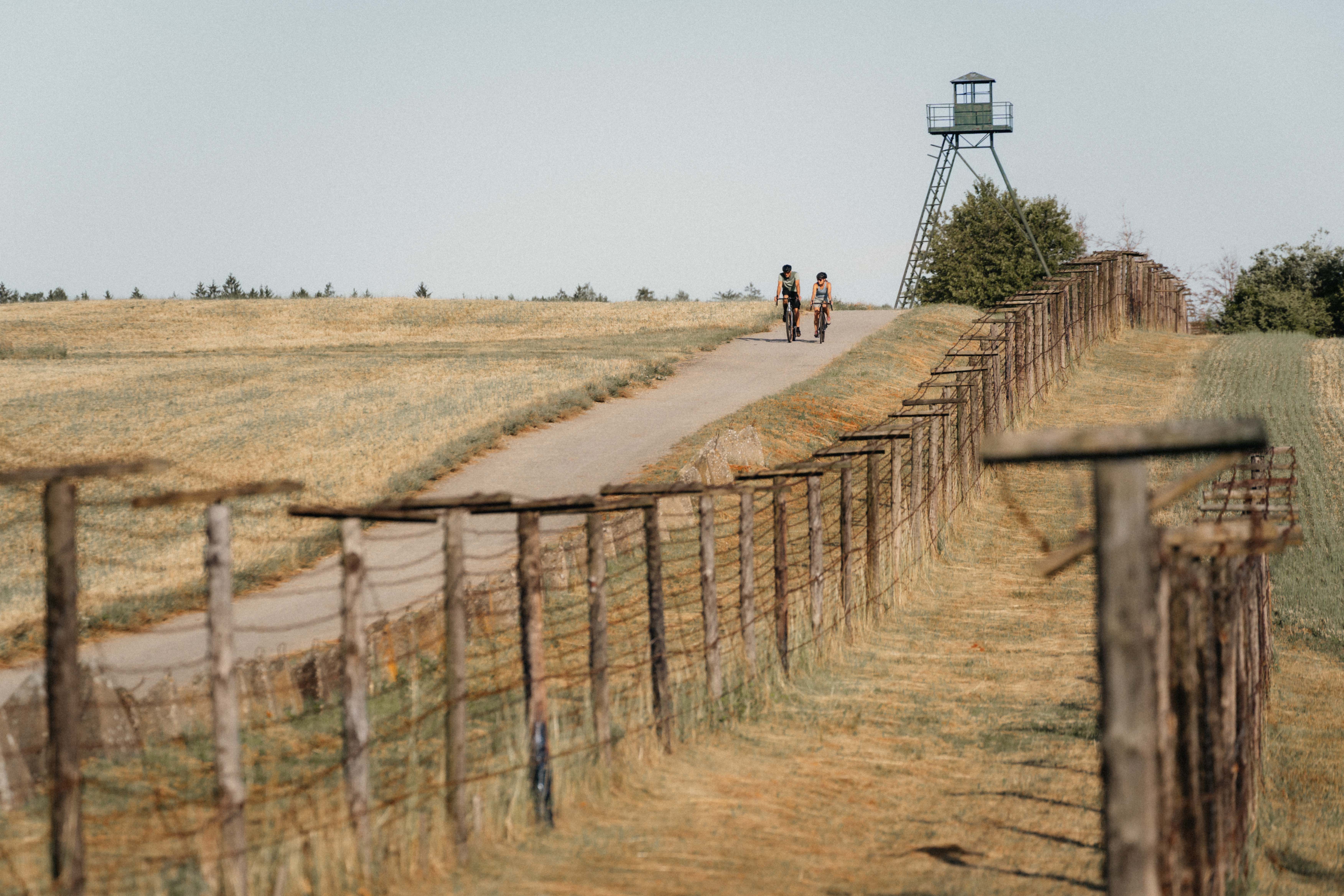 Two cyclists on a path next to an old border fence and a watchtower.