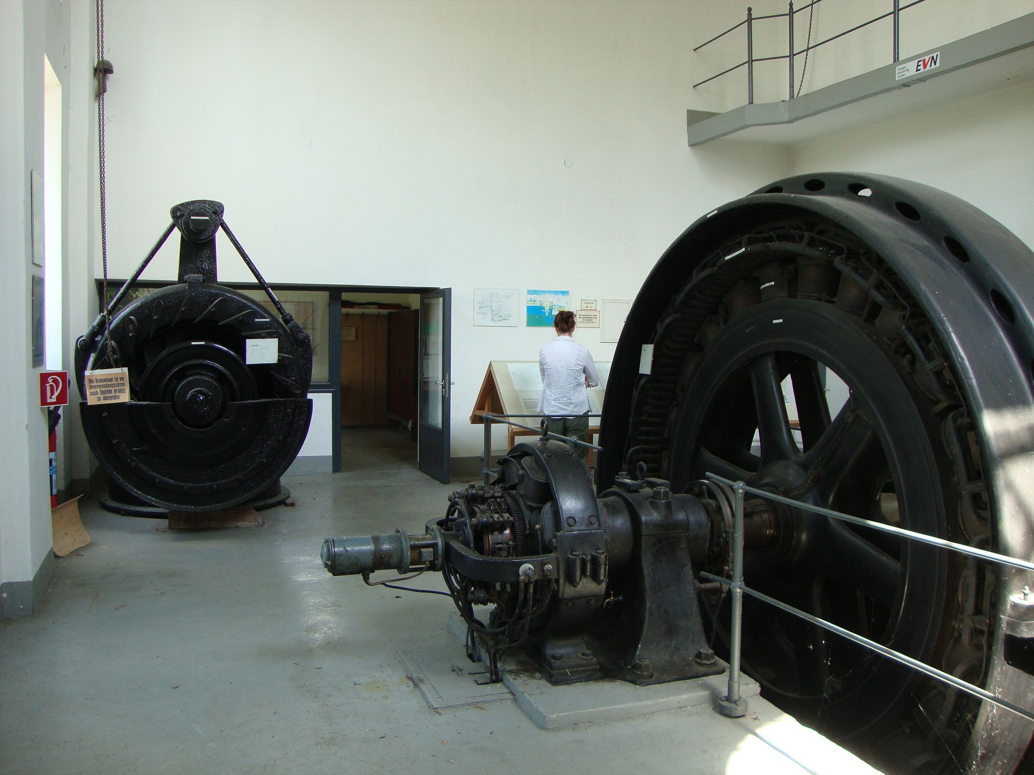 Interior view of a show power plant with large turbines and a person reading an information board.