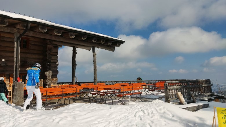 Snow-covered terrace of the Stoa Alm with wooden benches and tables, blue sky and clouds in the background.