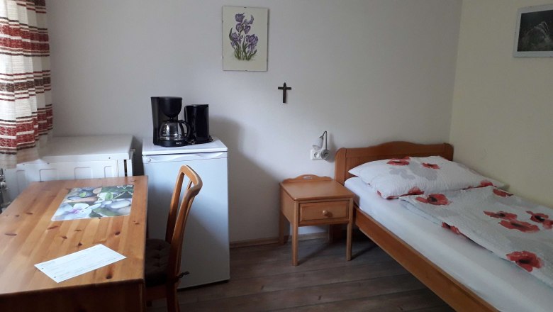 A simply furnished room with a bed, table, chair, fridge and coffee machine.